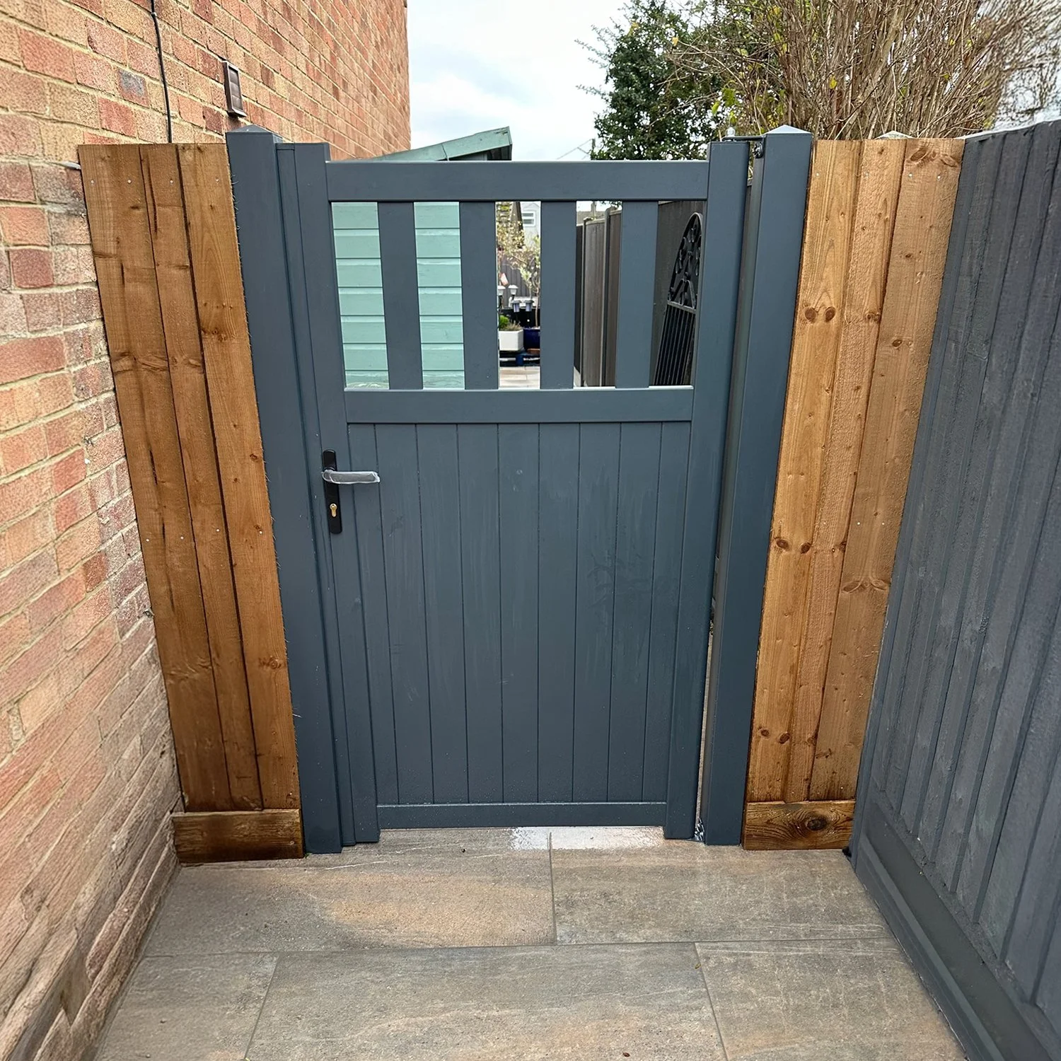 A blue wooden gate with a black handle, surrounded by a wooden fence on one side and a brick wall on the other, leading to an outdoor area.