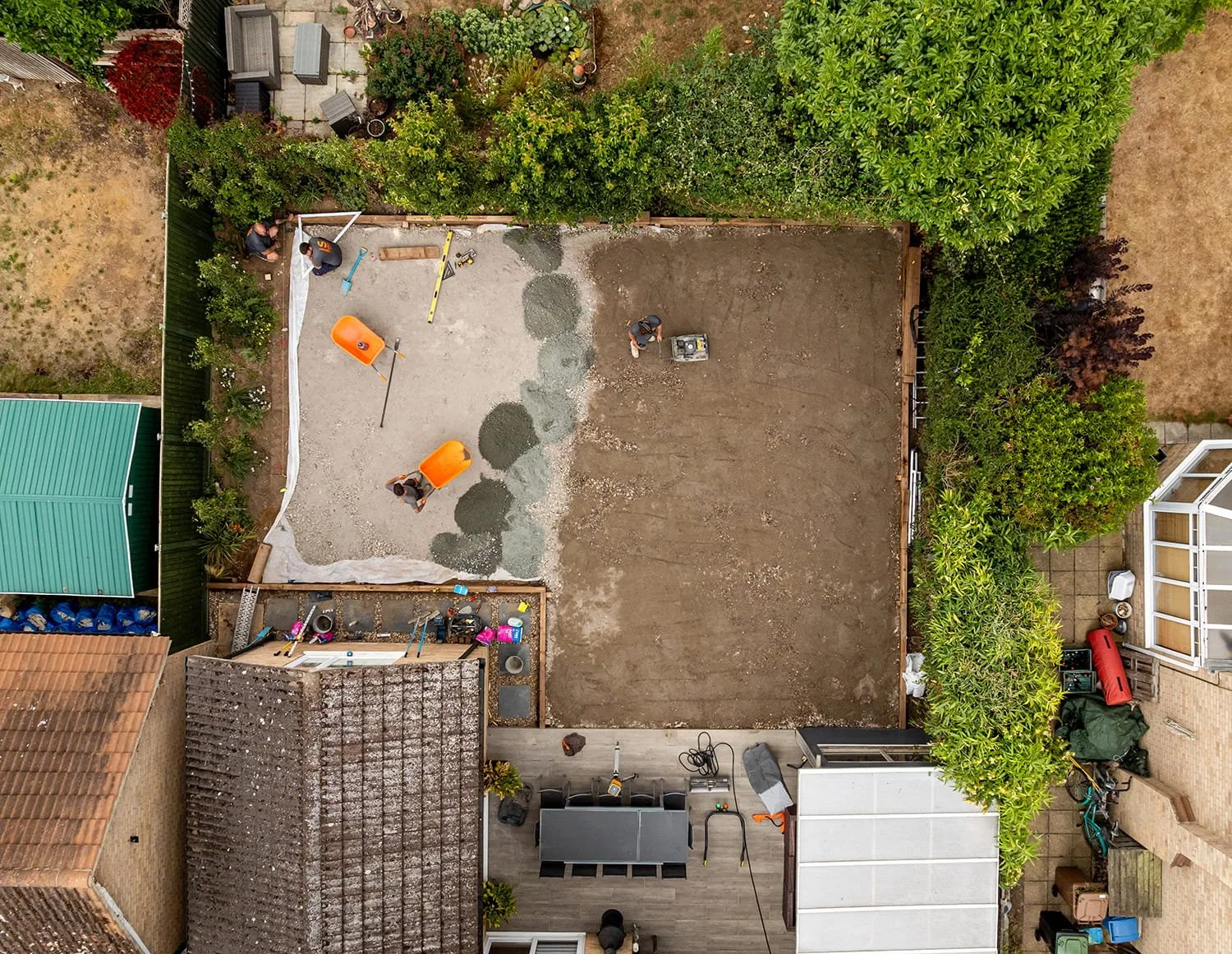 Backyard with two sections: one with a gravel surface being leveled by workers, and the other with bare soil. Construction tools and orange wheelbarrows are visible. Surrounding plants and trees border the yard, with neighboring houses and a patio area also visible.