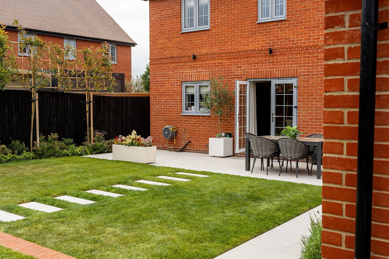 Backyard patio area with a table and chairs, potted plants, a garden with flowers, young trees, a black fence, and a red brick house with open glass doors. The yard has a well-maintained lawn and stepping stones.