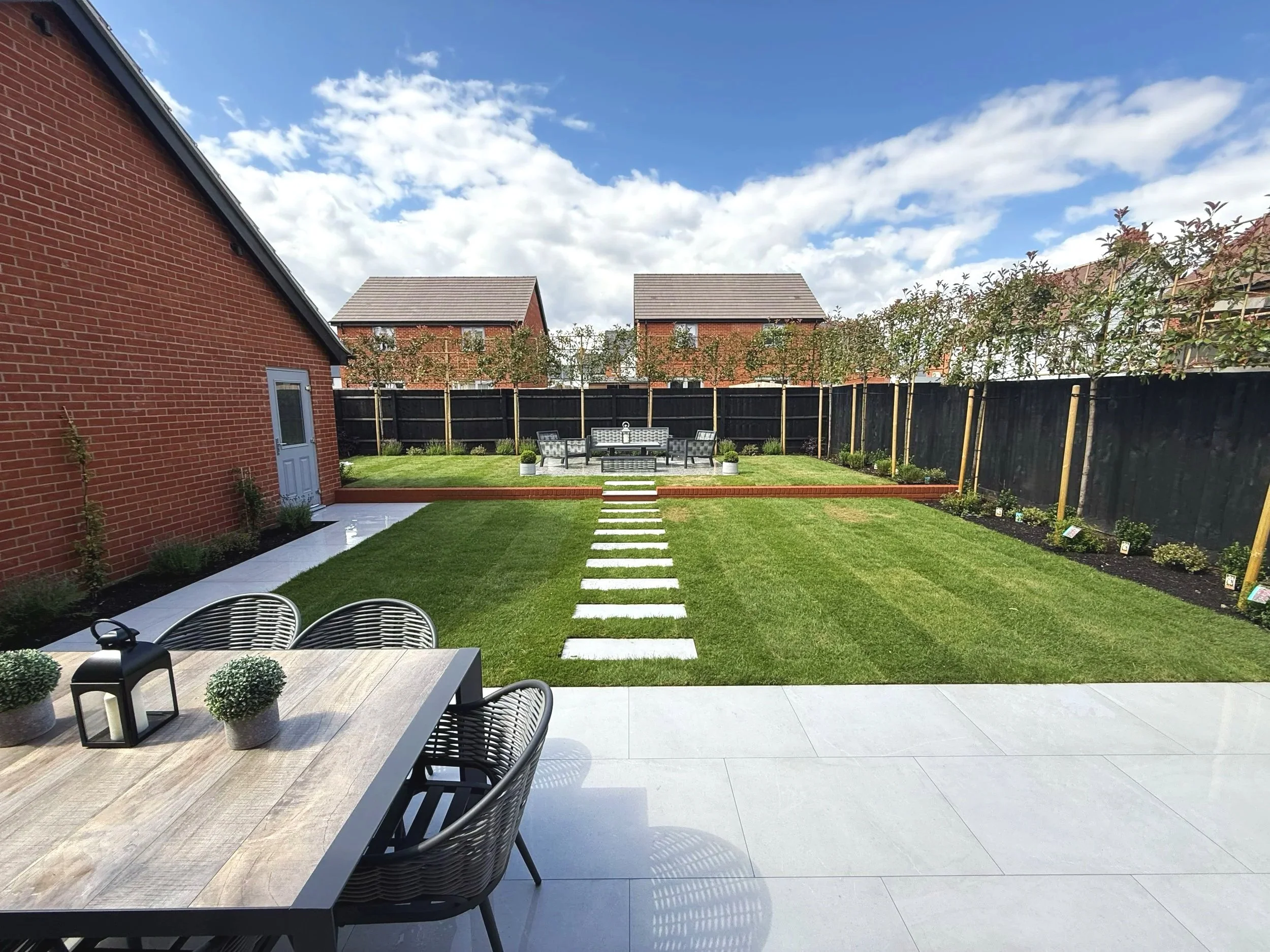 A backyard garden with a manicured lawn, stepping stones leading to a seating area with a bench, surrounded by a black fence and trees, under a partly cloudy sky.
