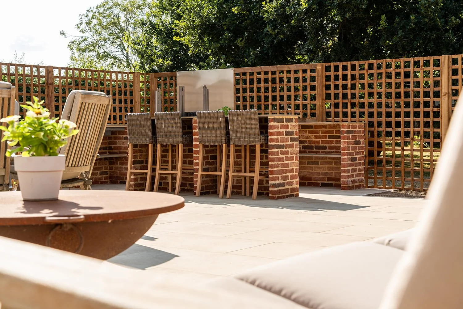 Outdoor patio with a brick bar counter and six wicker barstools, a potted plant on a wooden table, lounge chairs, a wooden lattice fence, and trees in the background.