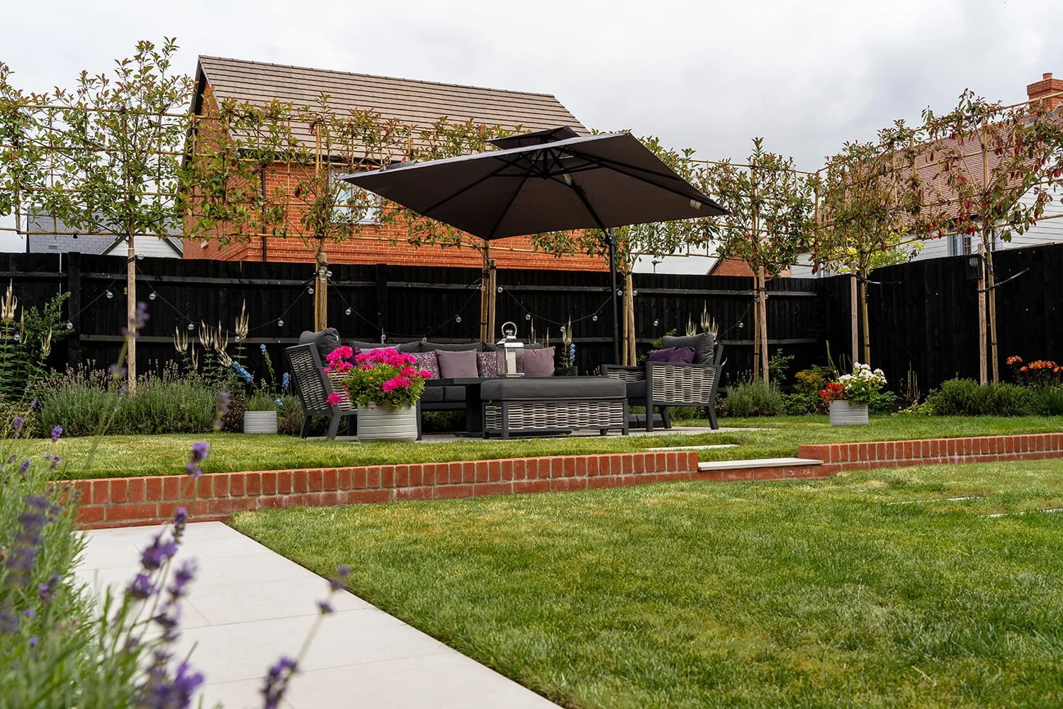 A backyard garden with a patio area featuring a black outdoor sofa, chairs, and a large black umbrella. There are potted plants with pink, white, and orange flowers, and a string of lights is hanging behind the seating area. The garden has a well-maintained lawn, a row of young trees with support stakes, and a black fence with brick accents.