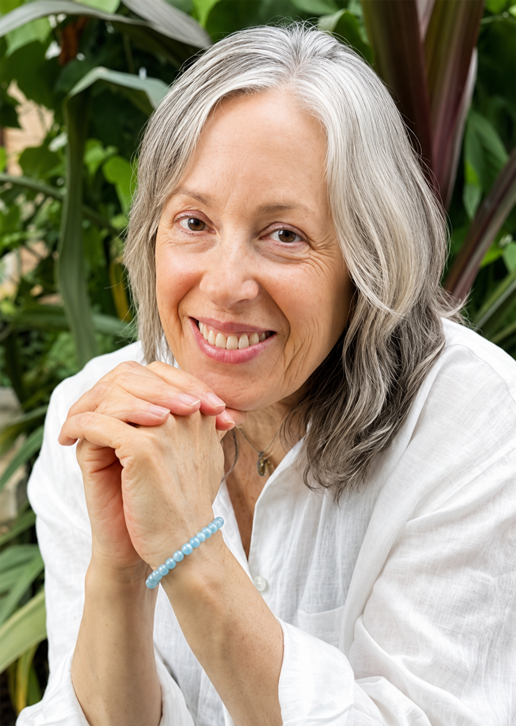 Close-up of a smiling elderly woman with gray hair, wearing a white shirt, with her hands clasped under her chin, sitting outdoors among green foliage.