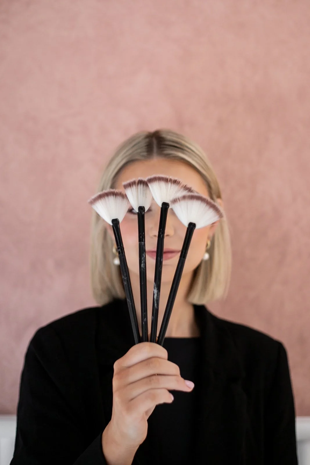 A woman with blonde hair holding five makeup brushes with white and brown bristles in front of her face, partially obscuring her face, against a pink background. Wedding and event makeup artist Mudgee