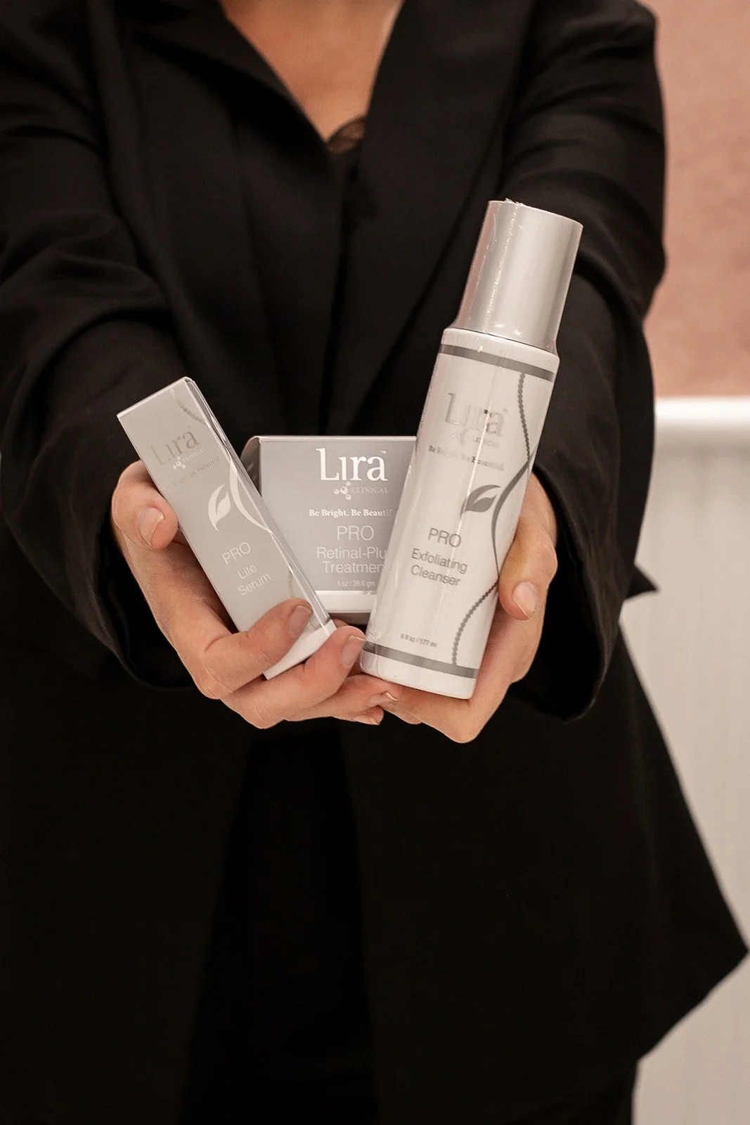 A person in a black blazer holding three skincare products from Lira Clinical, including a retinol-plump treatment, exfoliating cleanser, and lite serum. Lyra
