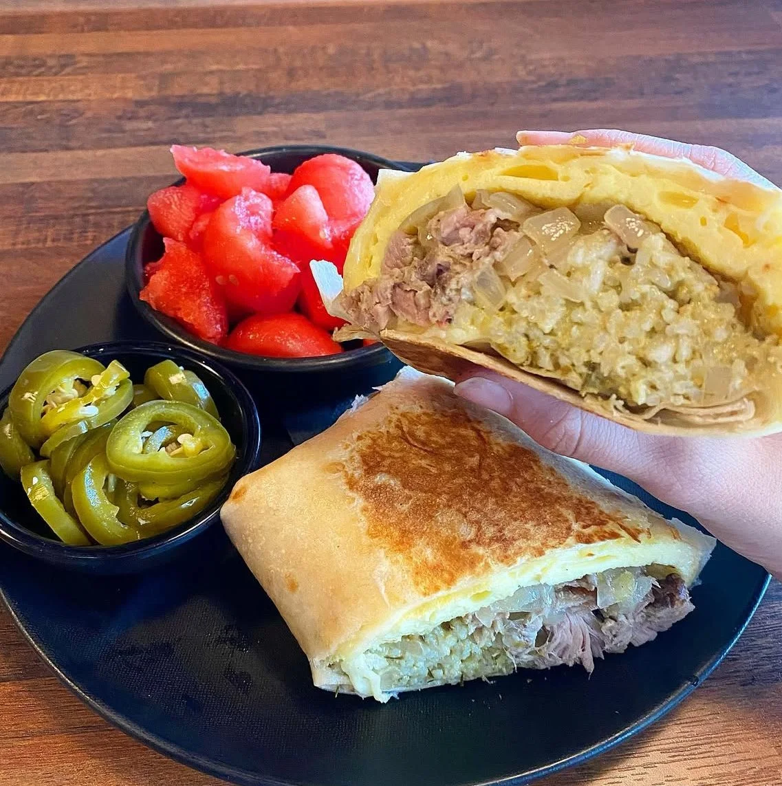 A plate of food with a breakfast burrito cut in half, stuffed with scrambled eggs, sausage, and onions, alongside slices of jalapenos, and small bowls of diced watermelon and sliced strawberries.