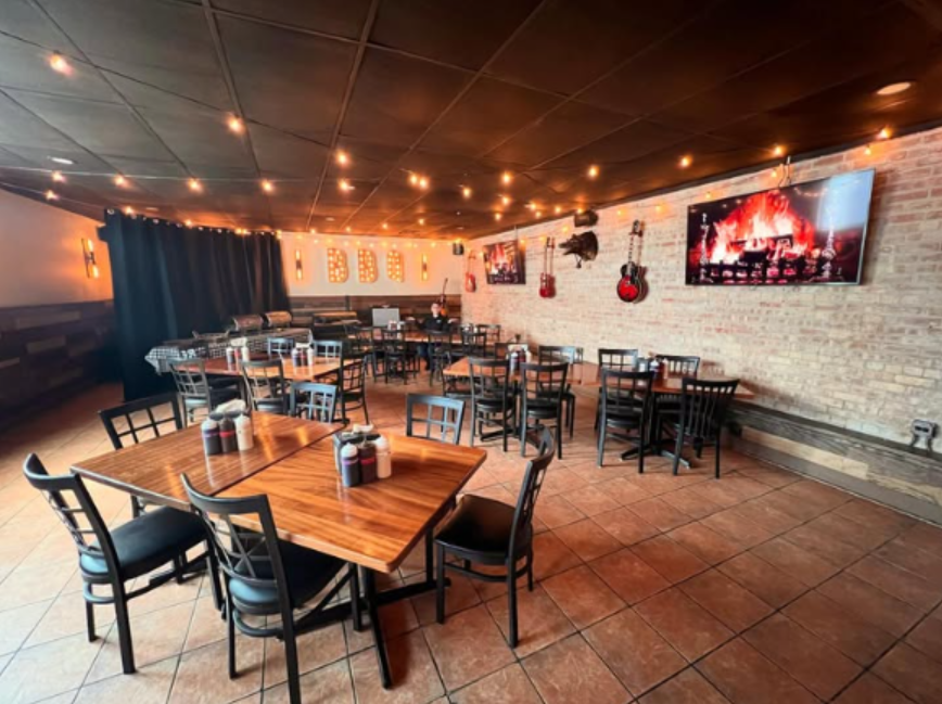 Empty restaurant with wooden tables and black chairs, string lights on ceiling, brick wall decorated with guitars and two TVs displaying flames, black curtains on one side.