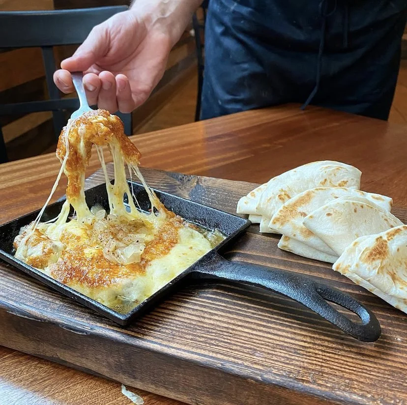 A person serving cheesy, baked dish with melted cheese and toppings from a cast iron skillet onto a plate, with folded flatbread on the side, on a wooden table.