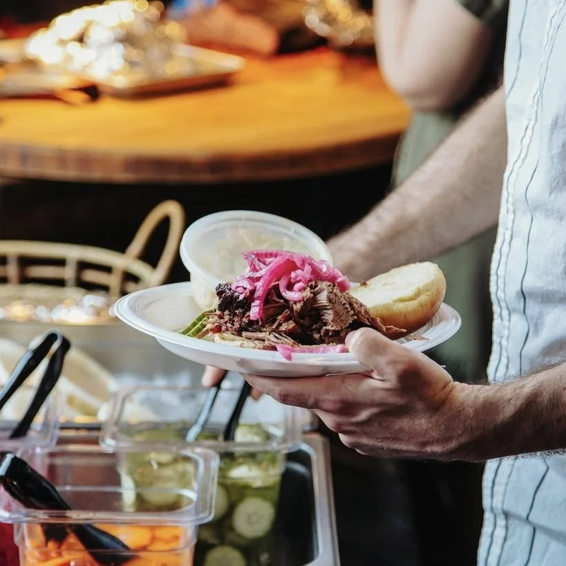 Person holding a paper plate with shredded beef, pickled onions, sliced bread, and a container of sauce, with a buffet of food and condiments in the background.