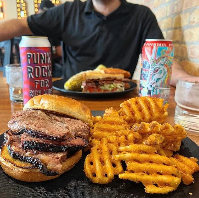 A plate of food with waffles, a burger, and waffle fries in front of a person with a drink, and another plate with a sandwich in the background.