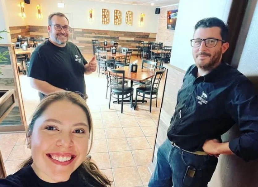 Three people in a restaurant or cafe, smiling for a selfie. One woman in the foreground, two men in the background near a wall with illuminated letters spelling 'EDBOX'.