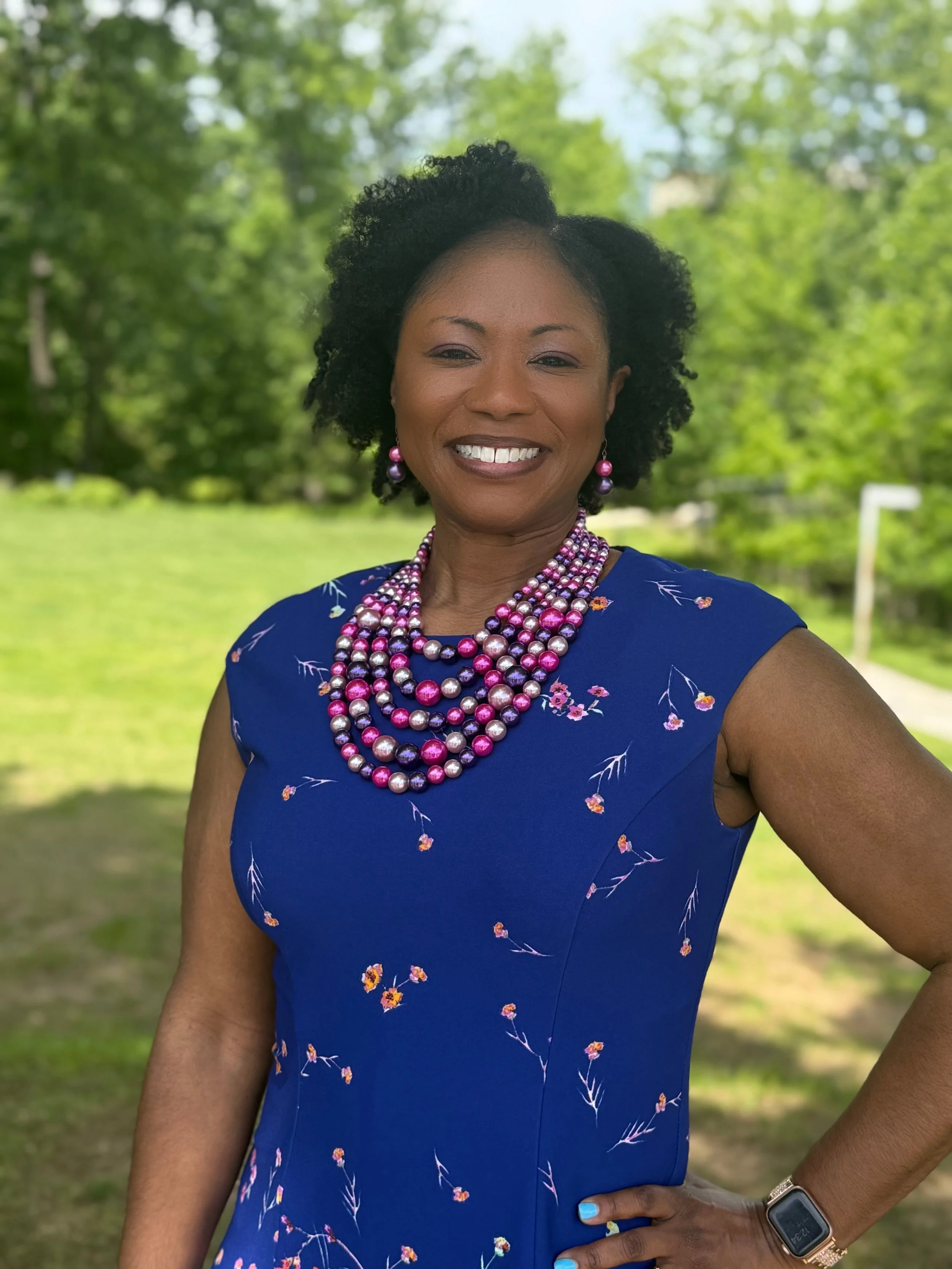 Dr. Petit- Woman smiling outdoors wearing a blue dress with pink and purple jewelry, standing in a green park.