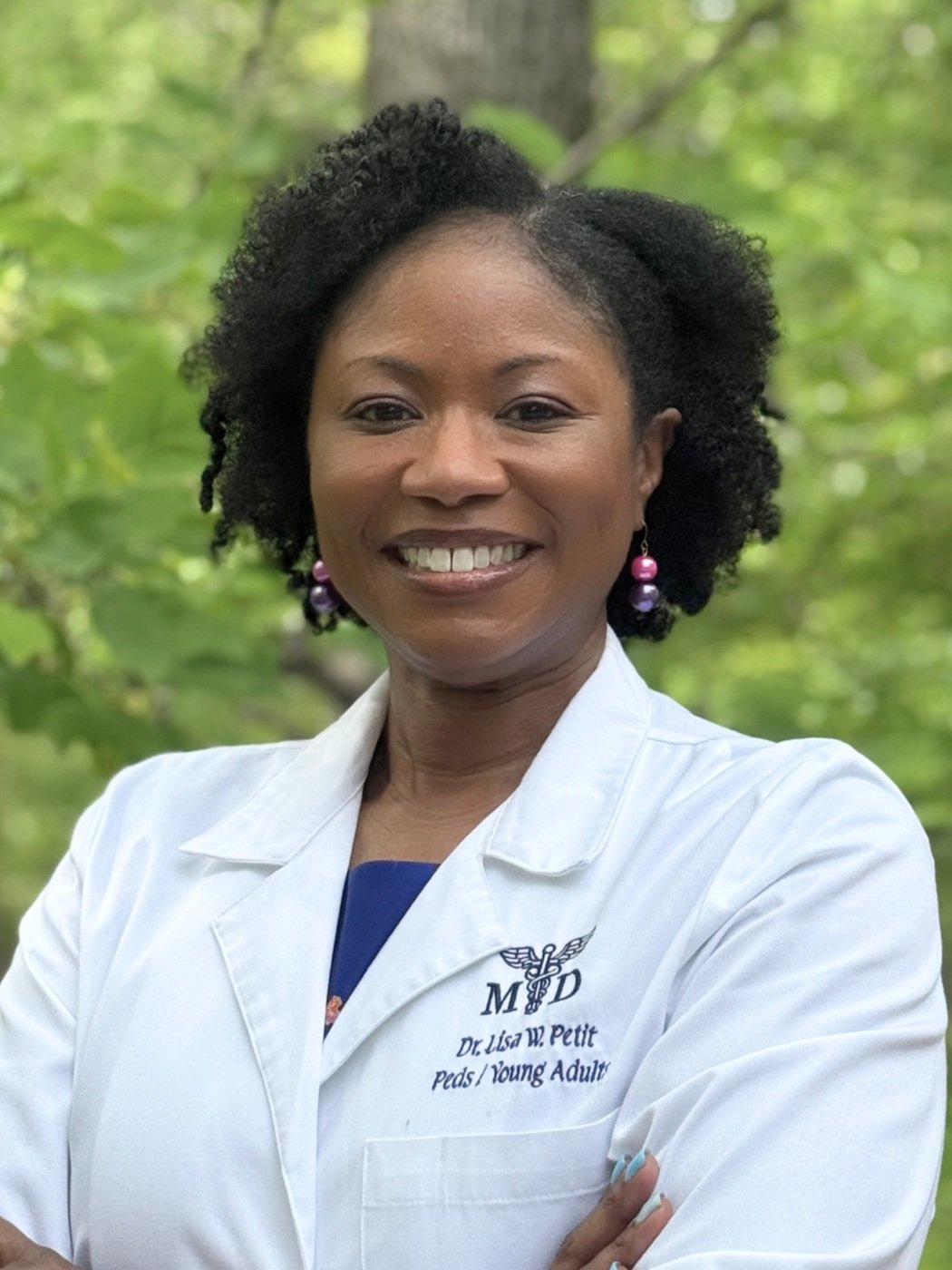 A woman with curly black hair and pink earrings, wearing a white coat with medical embroidery, standing outdoors with green foliage in the background.