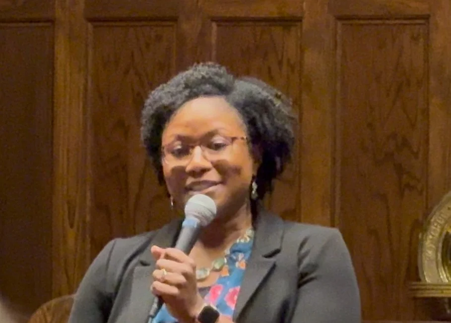 A woman with glasses and dark curly hair speaking into a microphone, standing in front of a wooden-paneled wall.