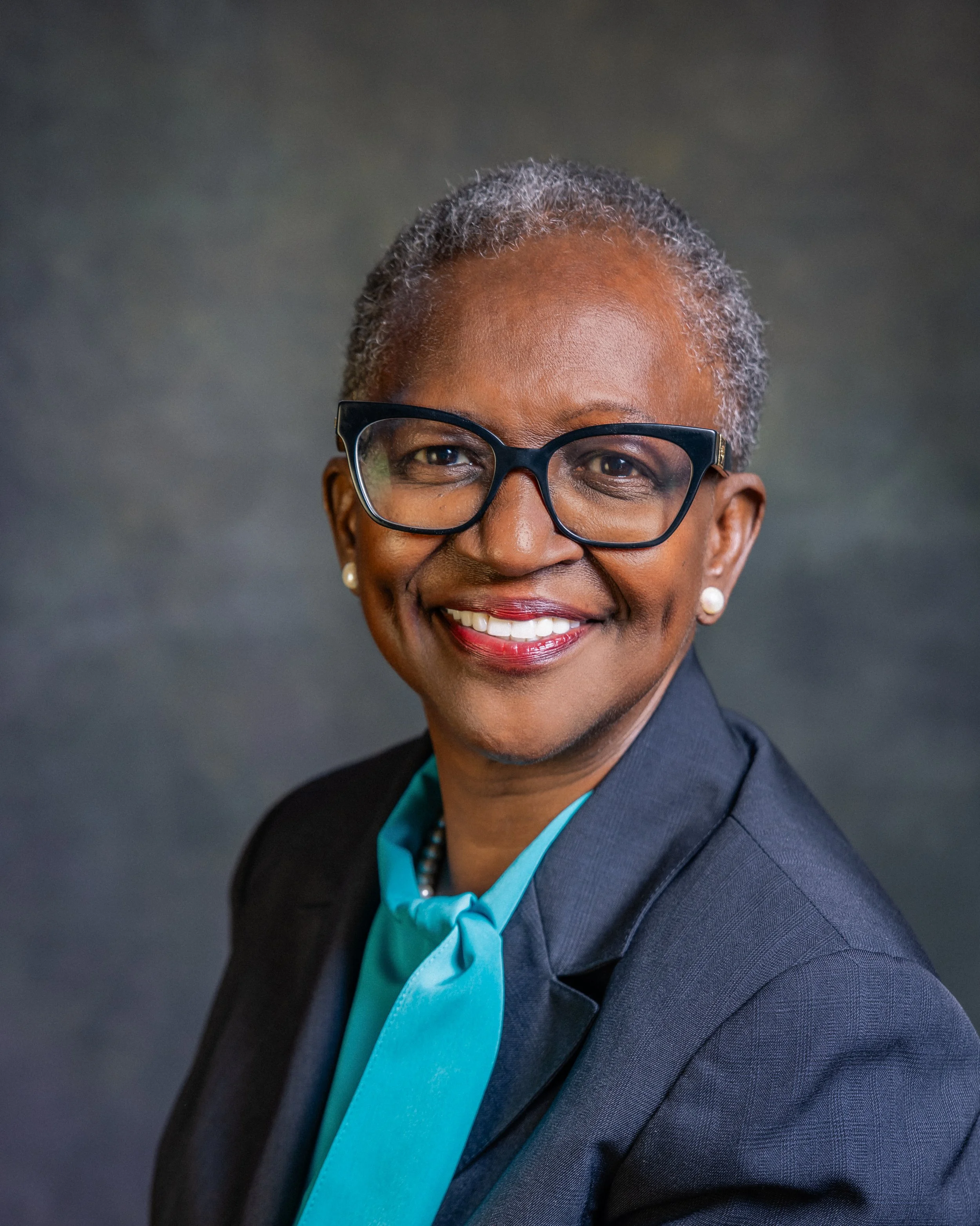 Portrait of a smiling Black woman with short gray hair, wearing glasses, pearl earrings, a teal blouse with a bow, and a dark blazer against a dark gray background.