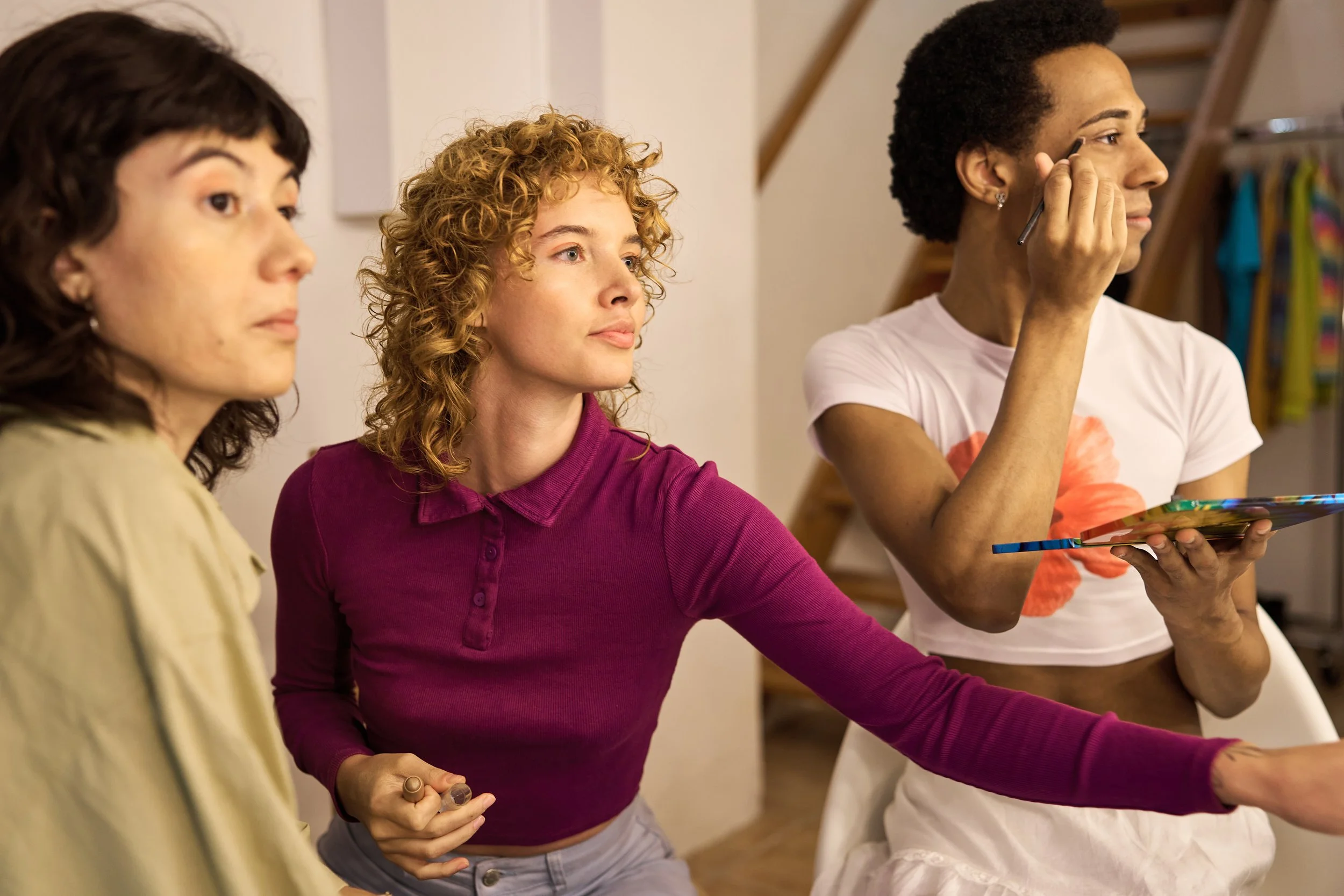 Three people standing in front of a mirror, with one person putting on makeup and holding a colorful book or folder, in a backstage dressing room.