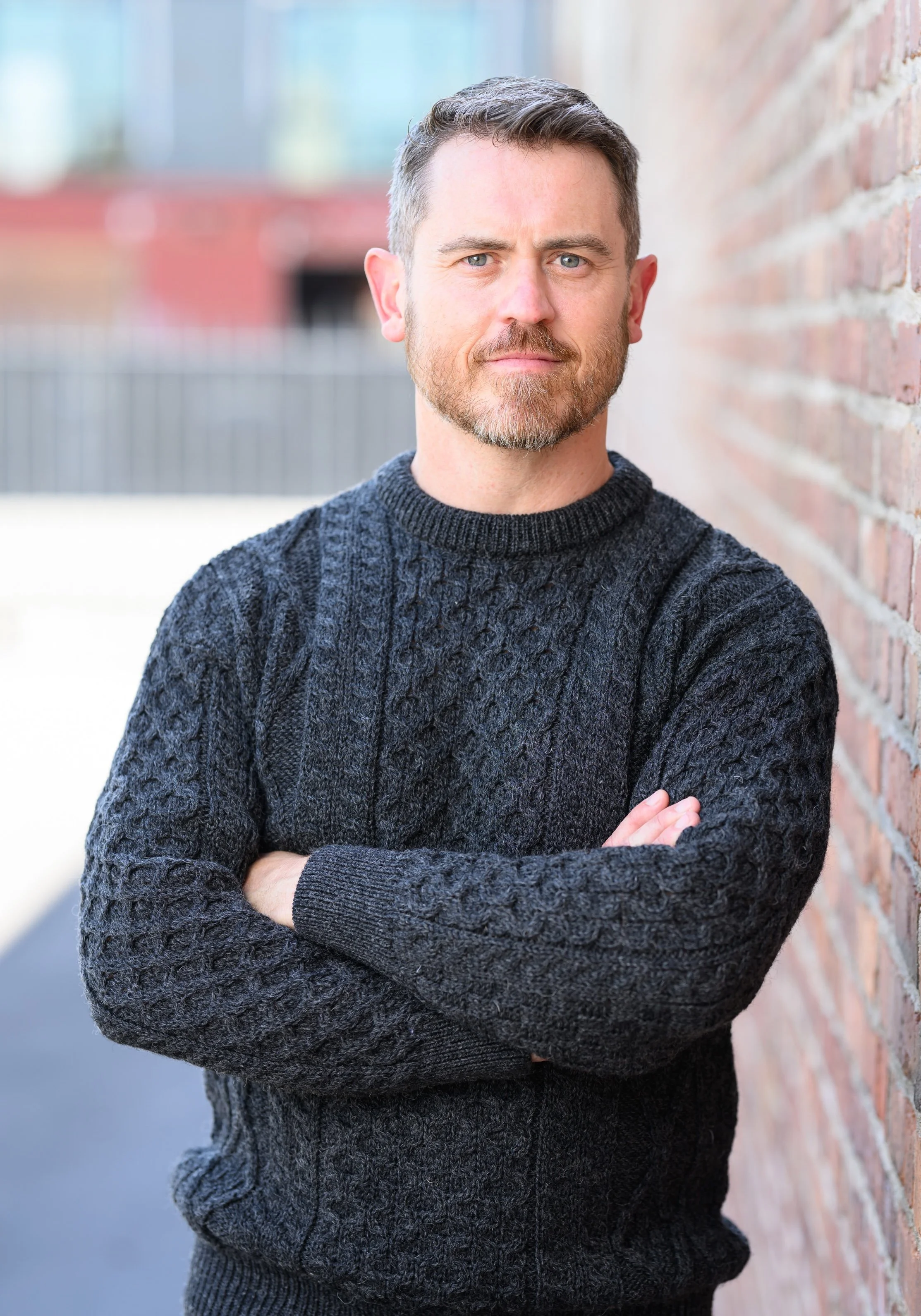 A man with short brown hair and a beard, wearing a black cable-knit sweater, standing outdoors with arms crossed, leaning against a brick wall.