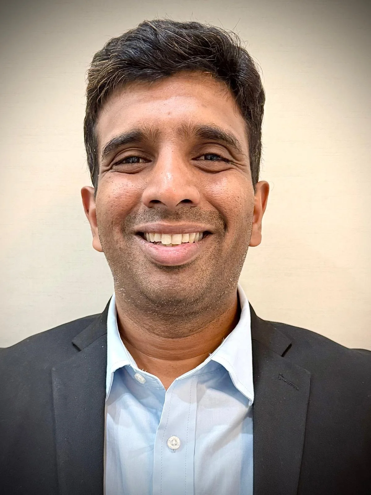 A smiling man in a dark suit and light blue dress shirt in front of a plain beige wall.