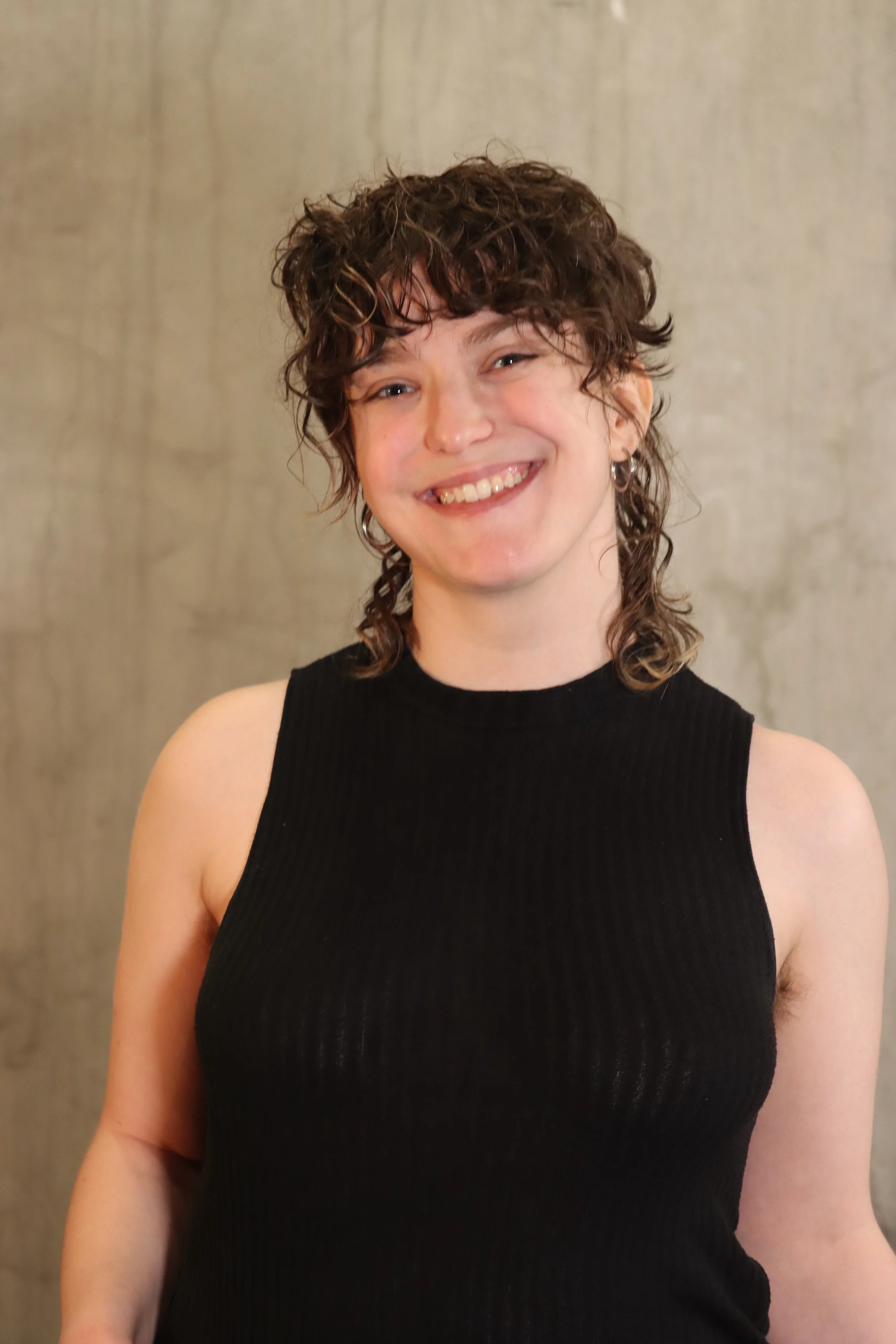 A woman with curly brown hair wearing a sleeveless black top, smiling against a plain light-colored background.