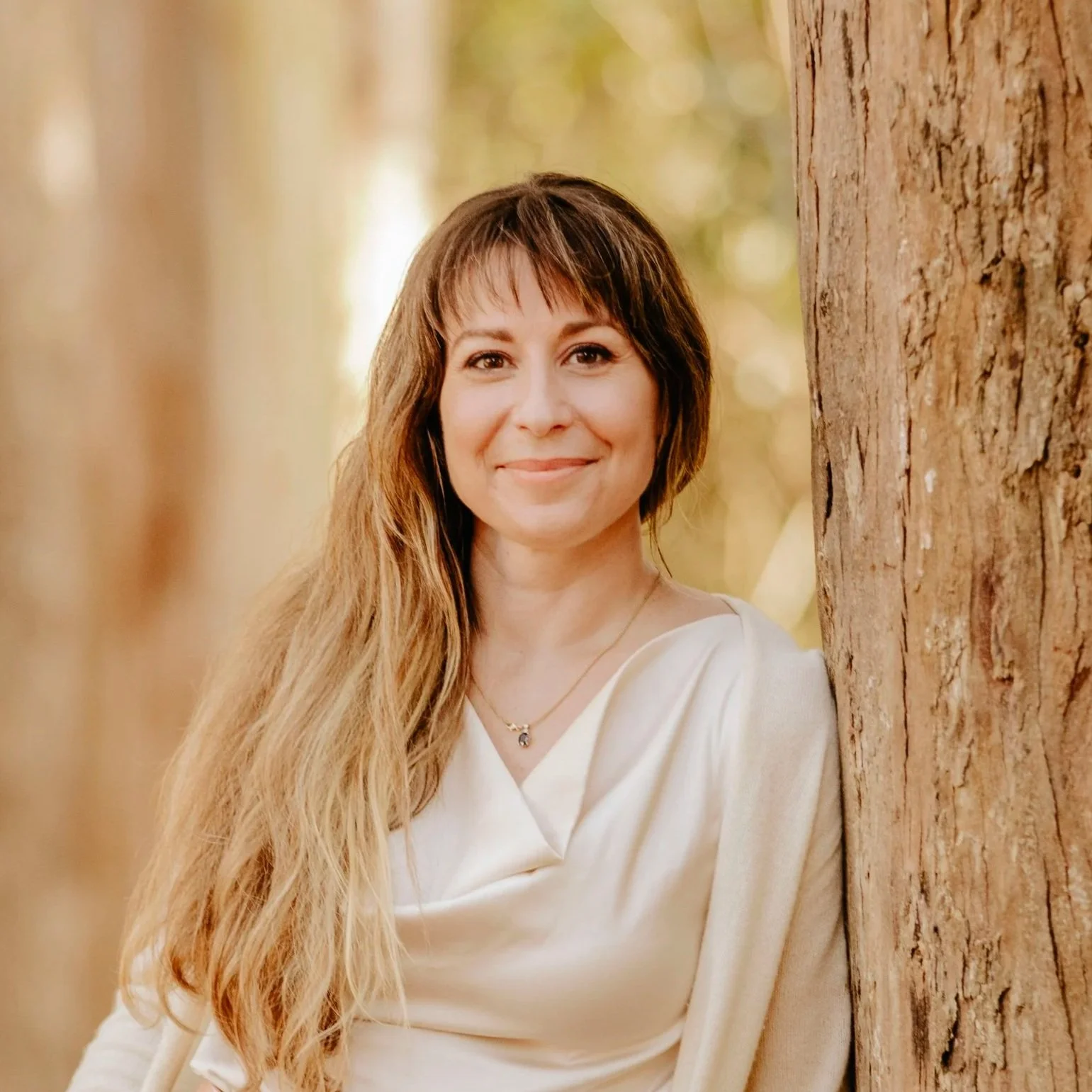 A woman with long, wavy hair standing outdoors next to a large tree with a blurred natural background.