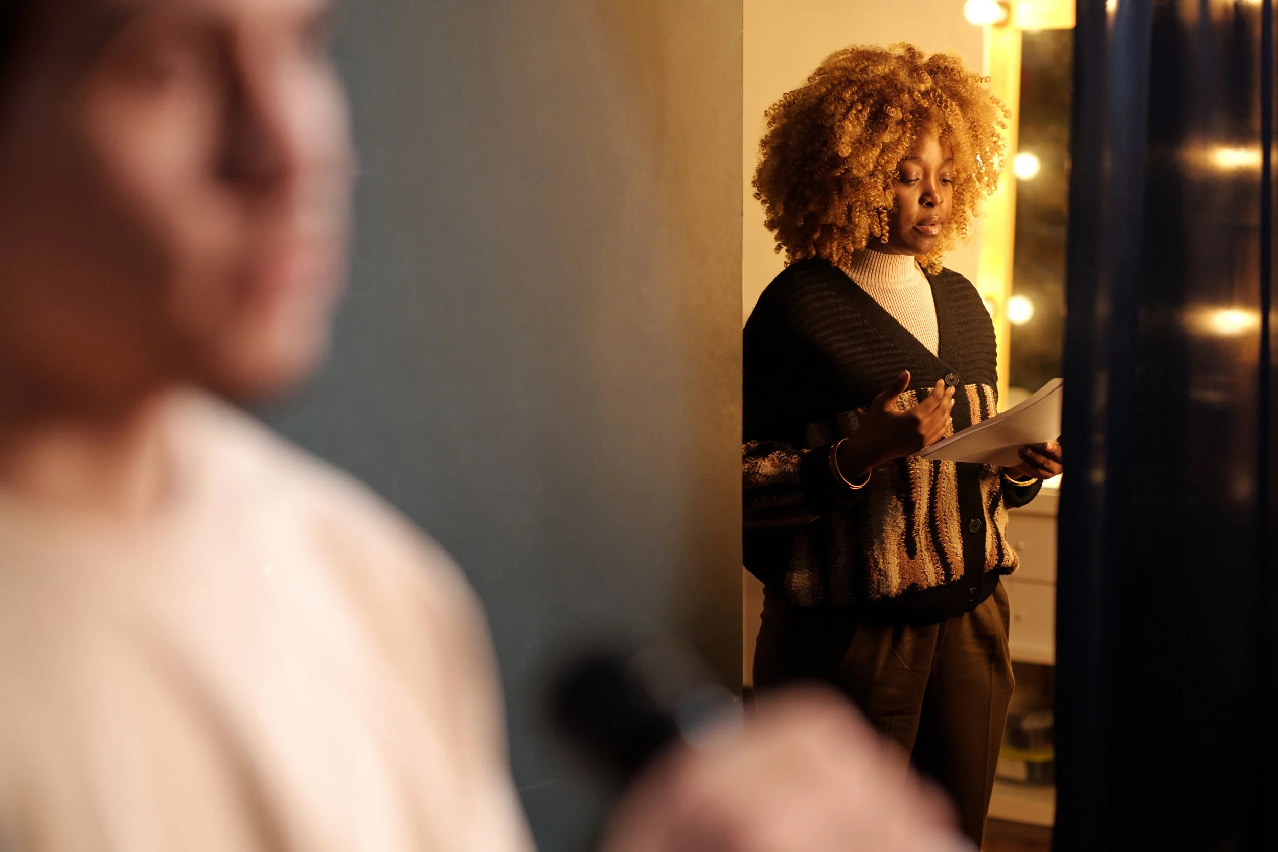 A woman with curly hair wearing a striped cardigan and holding a notepad stands reading to the side in a dimly lit room, with an out-of-focus person in the foreground holding a microphone.