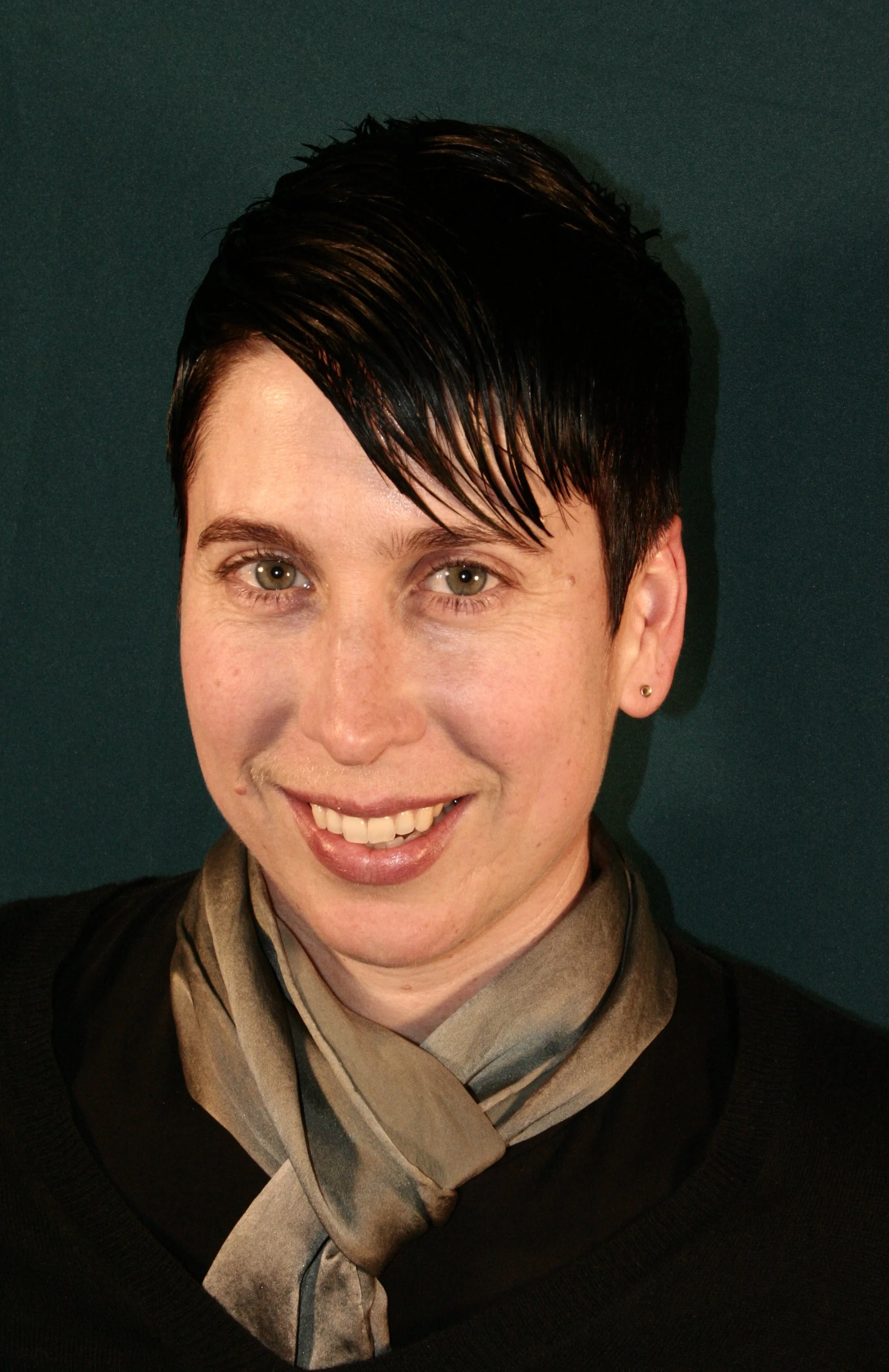 A woman with short dark hair, green eyes, wearing a silver silk scarf and a black top, smiling against a dark green background.