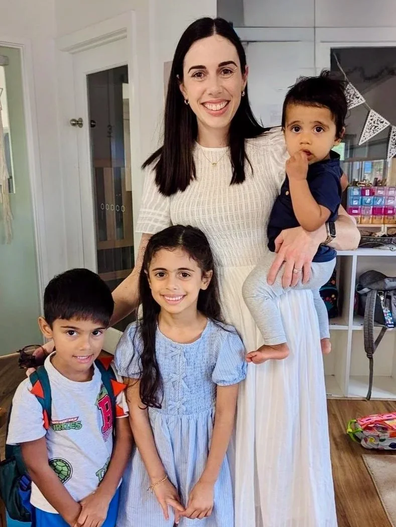 A smiling woman with long dark hair holding a young boy. Two children, a boy and a girl, stand in front of her in a room with shelves and a decorated wall in the background.