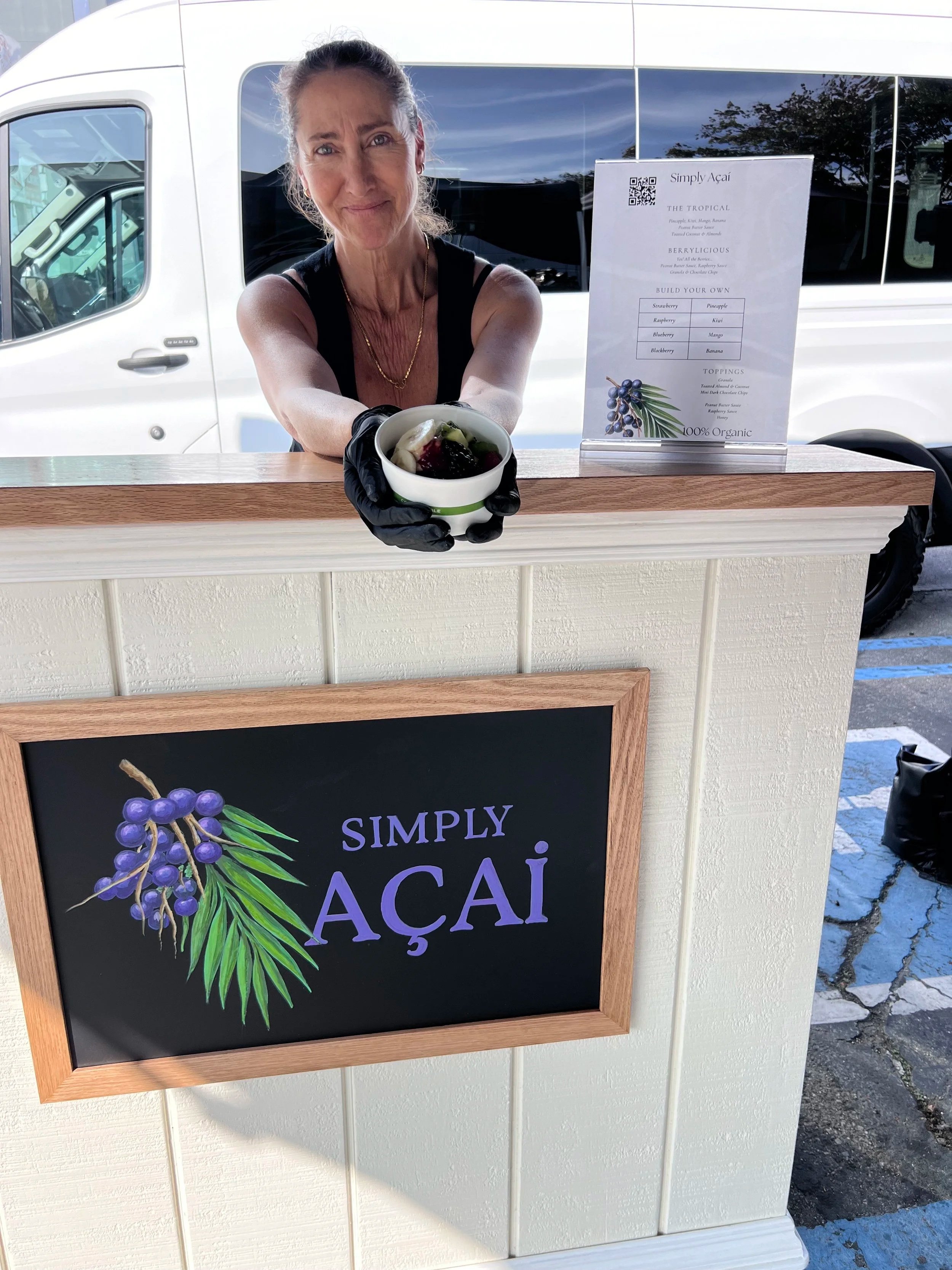 A woman holding a cup of açaí, standing behind a counter with a sign that shows a menu and ingredients for açaí bowls, outdoors with a white van and parking lot visible in the background.