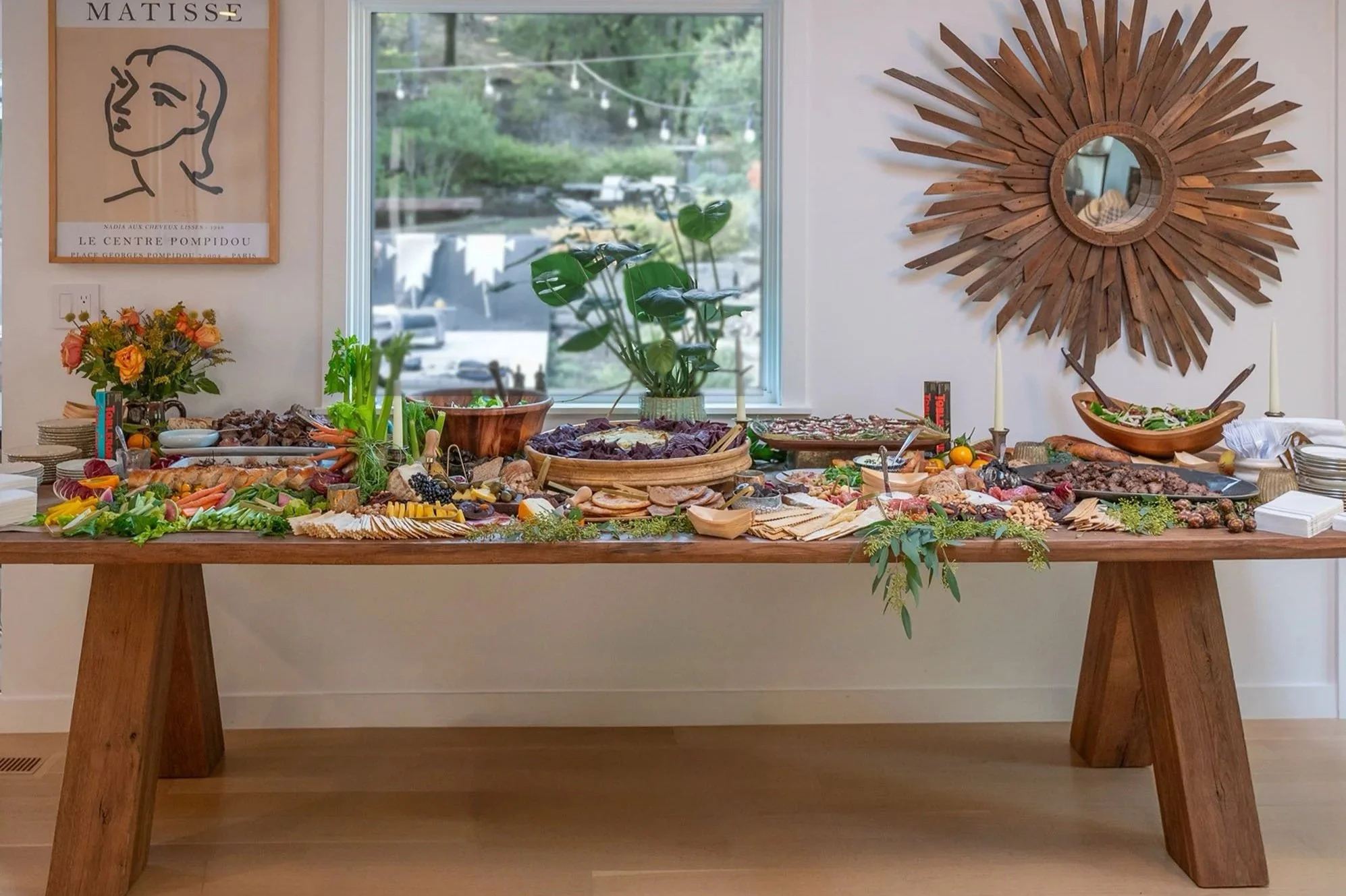 A buffet table filled with various dishes, vegetables, fruits, and charcuterie, set against a background of a window and a large decorative wooden starburst mirror on the wall.