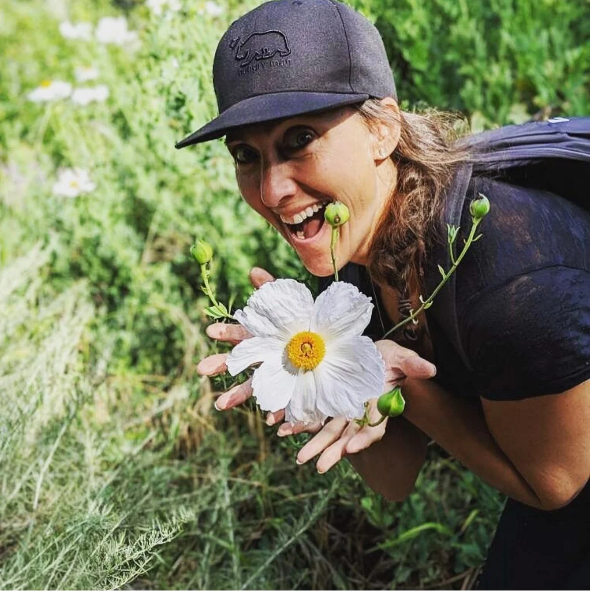A woman with a baseball cap and black shirt holds a white flower with a yellow center, with small green buds attached, near her face in a lush green outdoor setting.