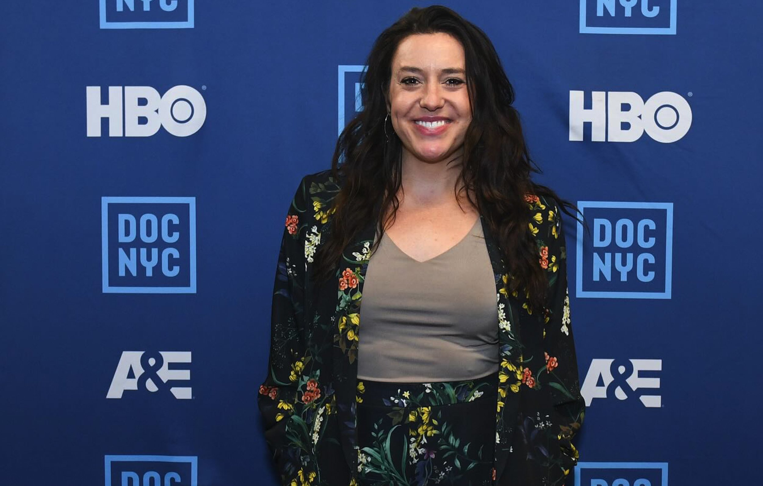 Janelle Draper with long dark hair smiling in front of a blue background featuring logos of HBO, DOC NYC, and A&E, wearing a black floral blazer over a beige shirt.