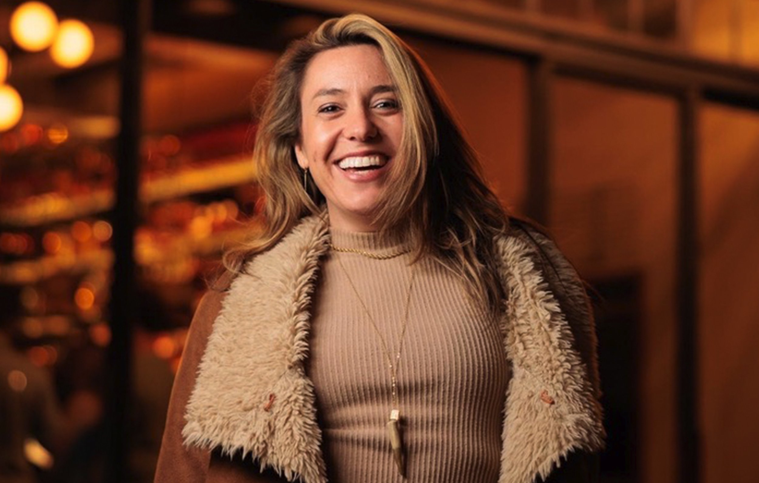 A young woman with long wavy hair smiling in a warmly lit indoor space, wearing a beige top, a brown jacket with a shearling collar, and layered necklaces.