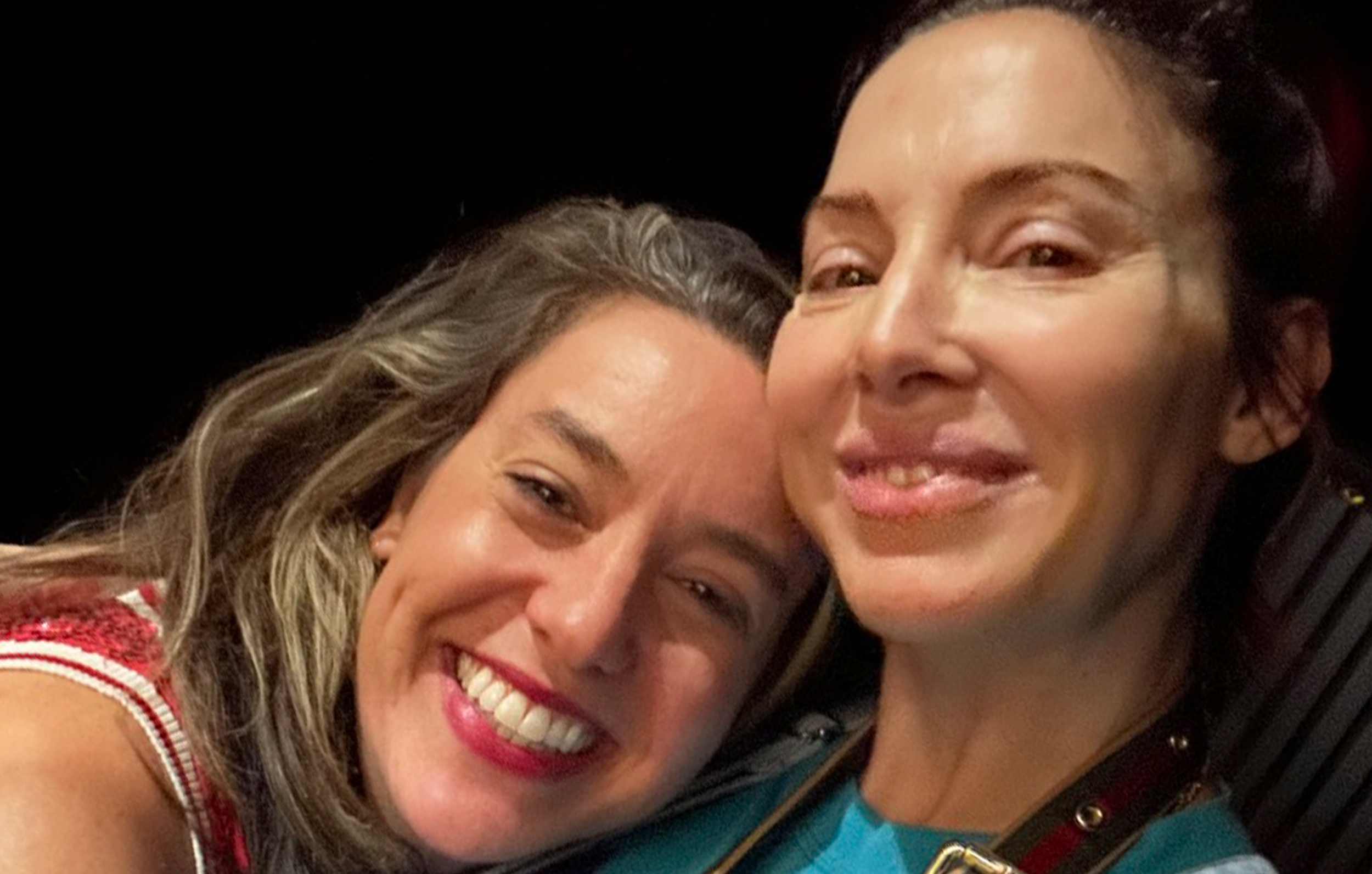 Two women smiling and leaning close together, one with long wavy blonde hair and the other with short dark hair, both in close-up portraits.
