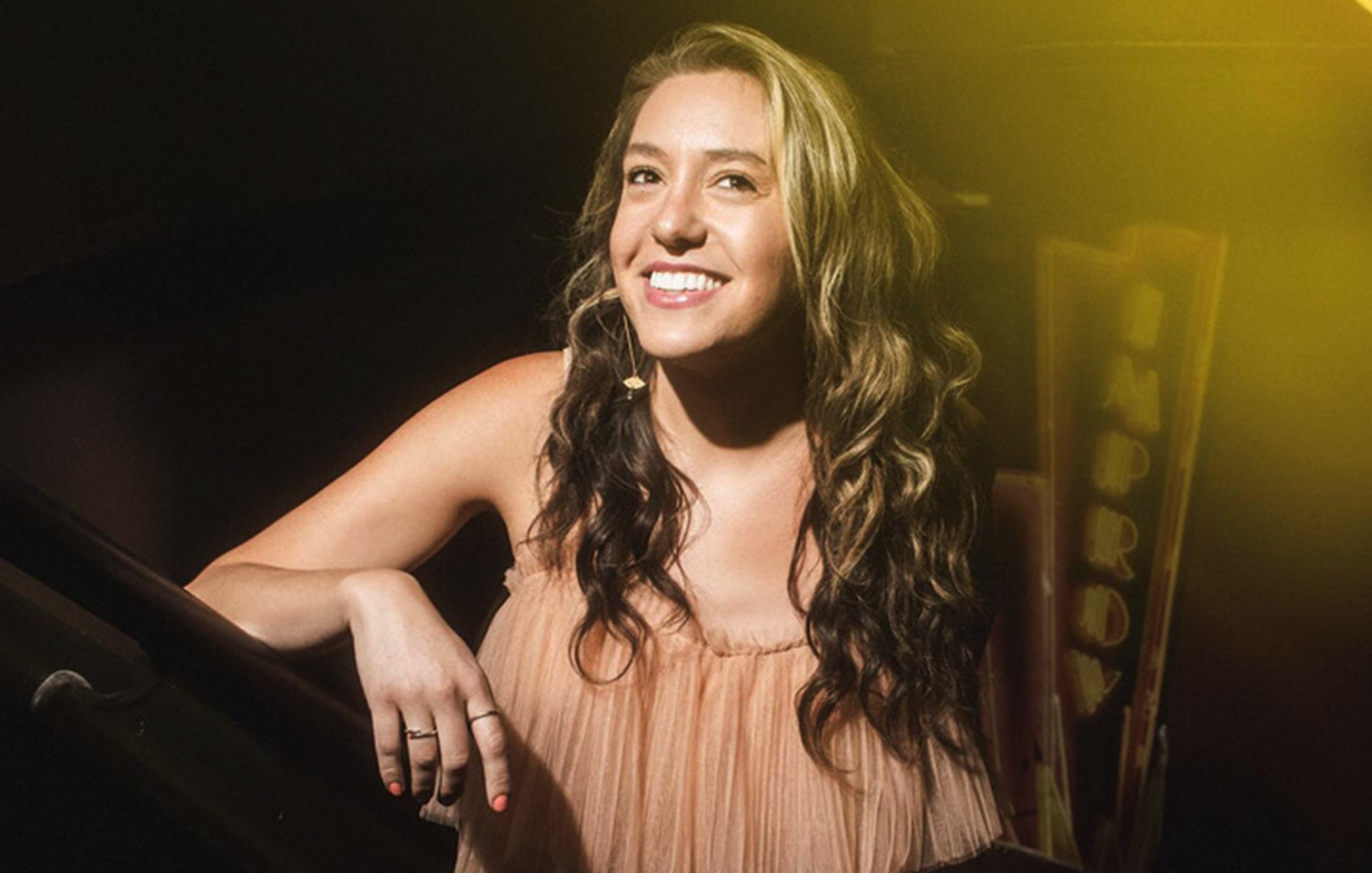 A smiling young woman with long, curly hair, wearing a beige dress, posed by a piano with a background featuring colorful, illuminated letters.