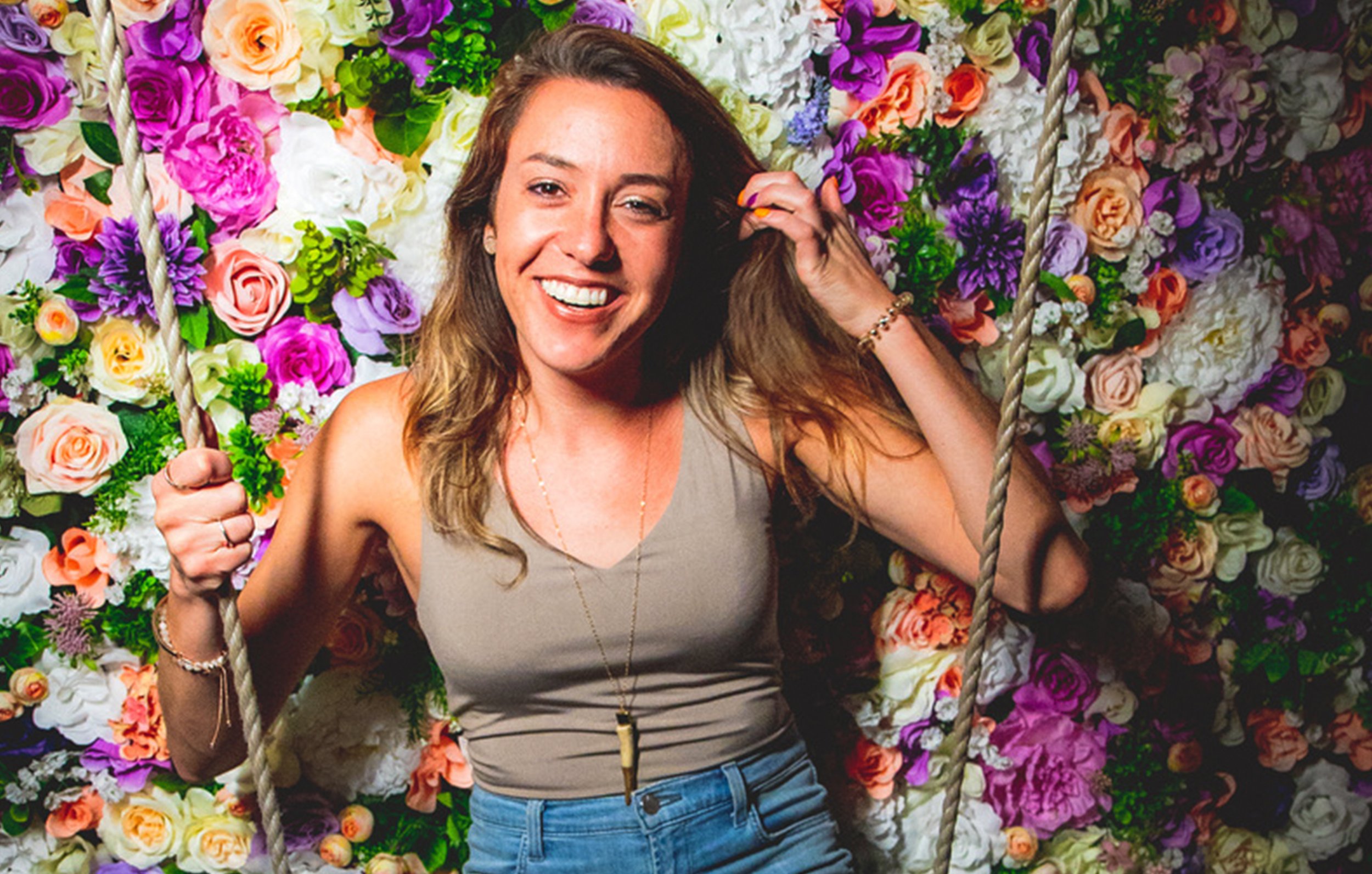 A smiling woman with wavy brown hair sitting on a swing in front of a background filled with colorful flowers, wearing a beige sleeveless top and jewelry.
