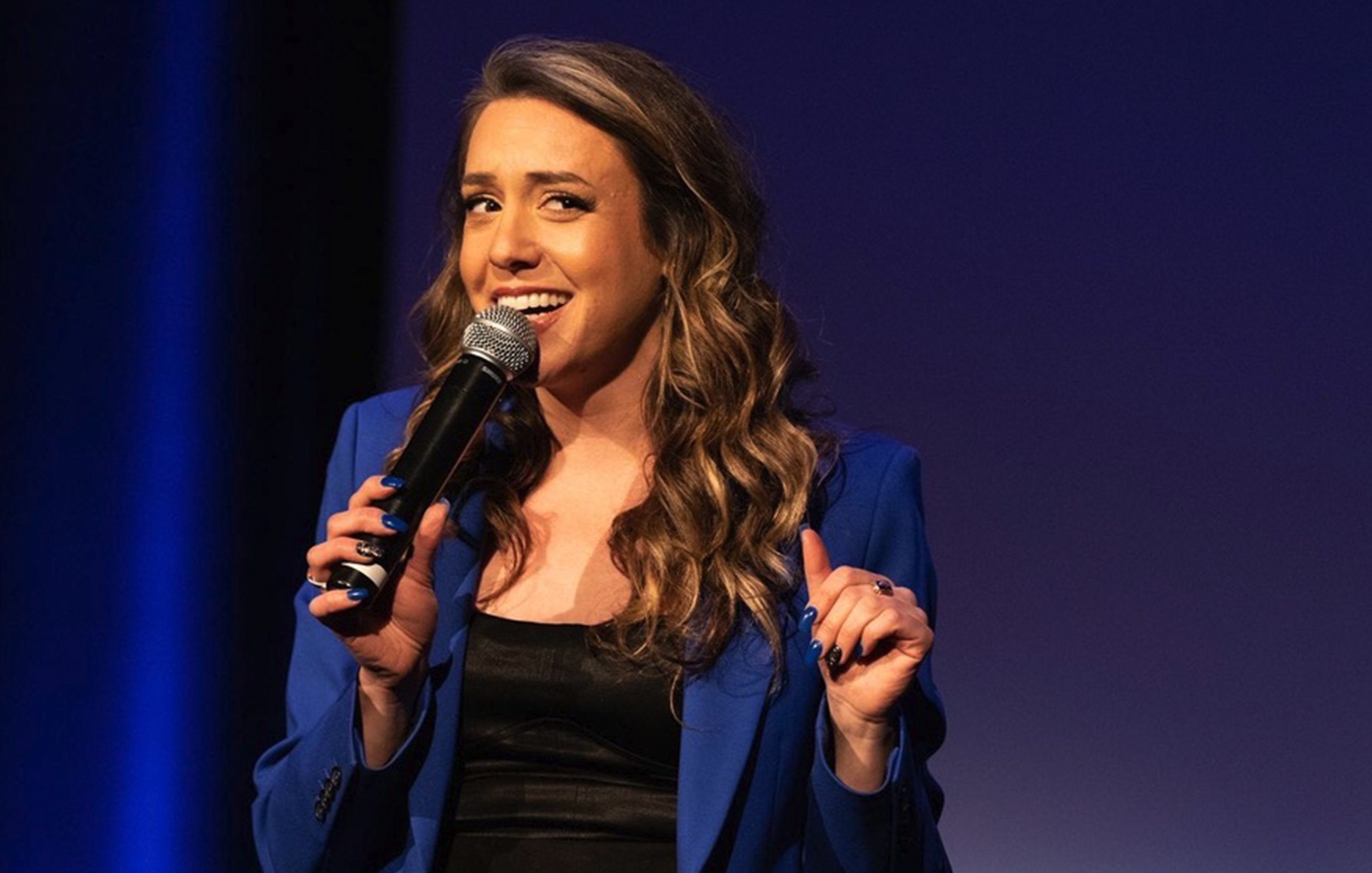Janelle Draper wearing a blue blazer holding a microphone on stage. She is smiling and making a point with her other hand.