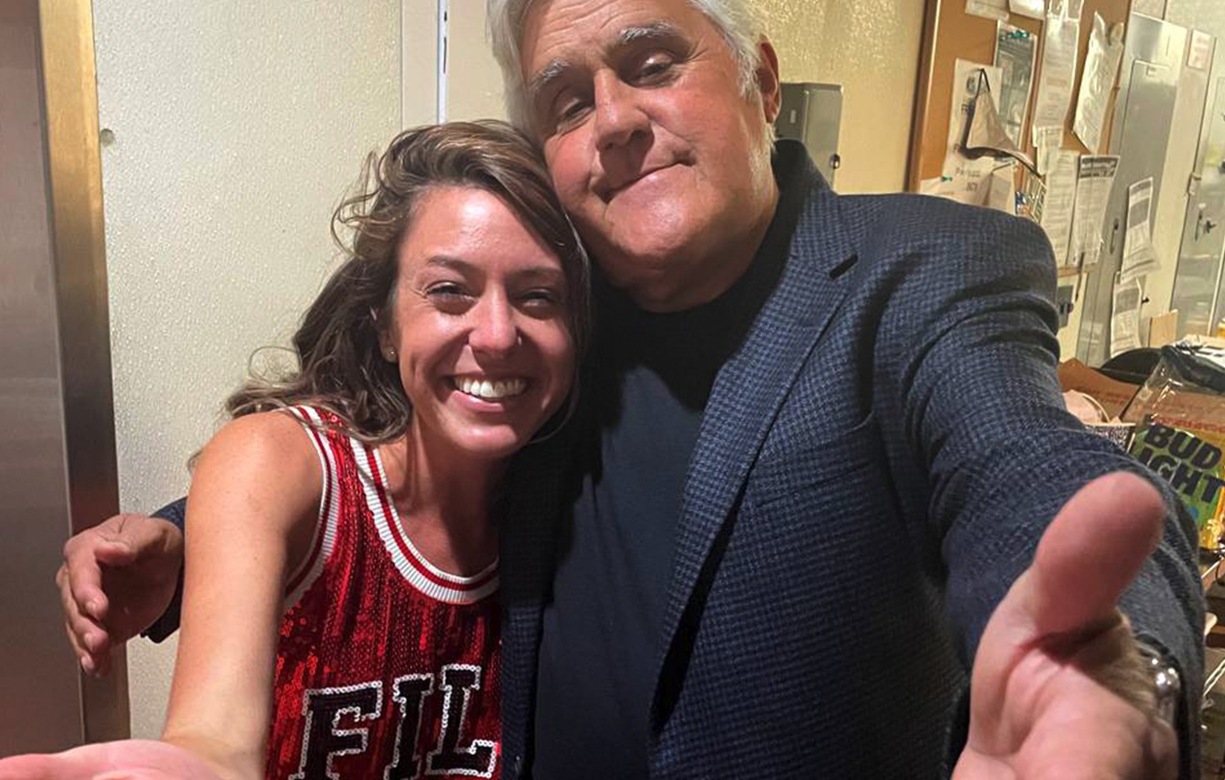 Janelle Draper and a man are taking a selfie together, both smiling. The woman has brown hair and is wearing a red sports jersey. The man has gray hair, is wearing a dark blazer and has his arm extended to take the photo.