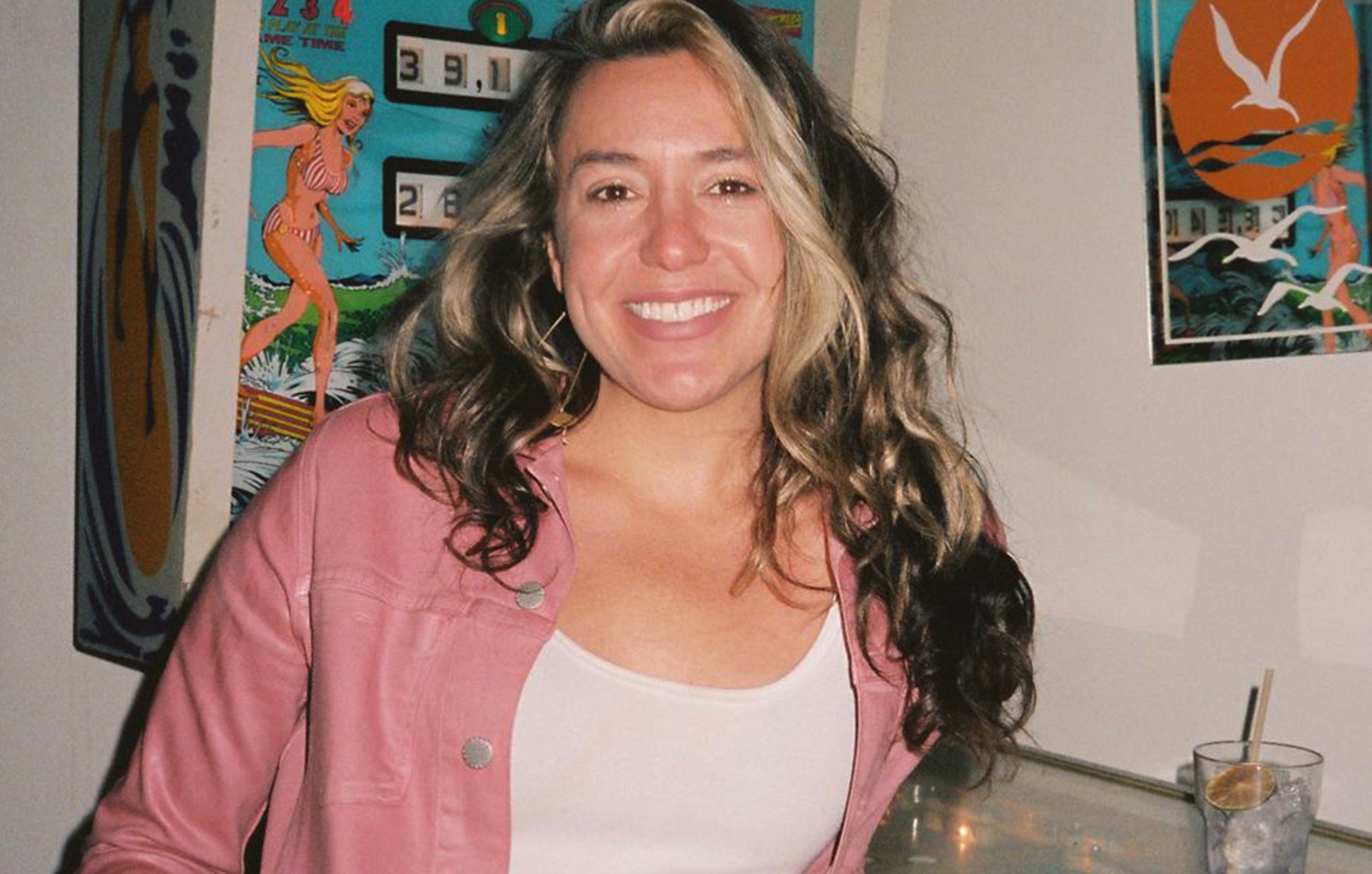 A woman with long, curly hair and a bright smile, wearing a light pink jacket over a white top, standing indoors with colorful posters and artwork on the wall behind her.