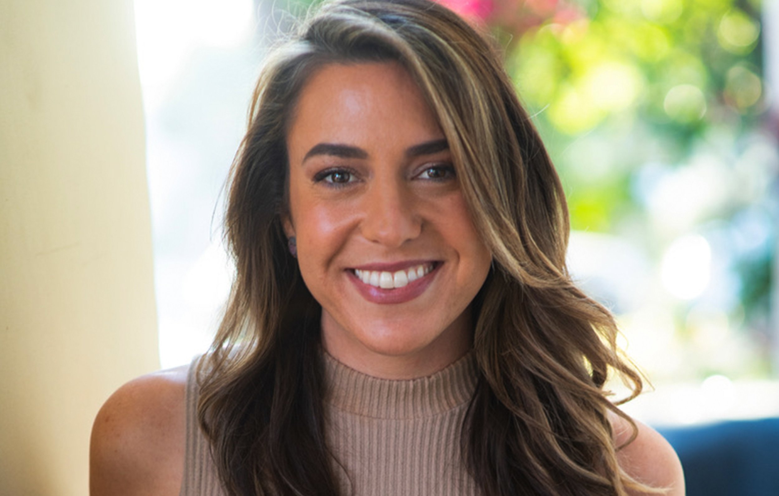 A smiling woman with long, wavy brown hair, wearing a beige sleeveless top, is indoors with a bright, colorful, blurred outdoor background.
