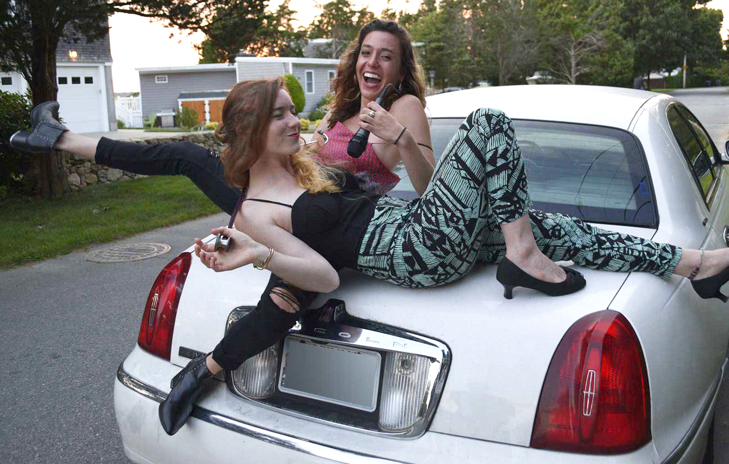 Two young women with curly hair posing on the hood of a silver police car parked on the street in front of residential houses, one woman is stretching her leg and holding a car key, the other is holding a microphone and smiling.