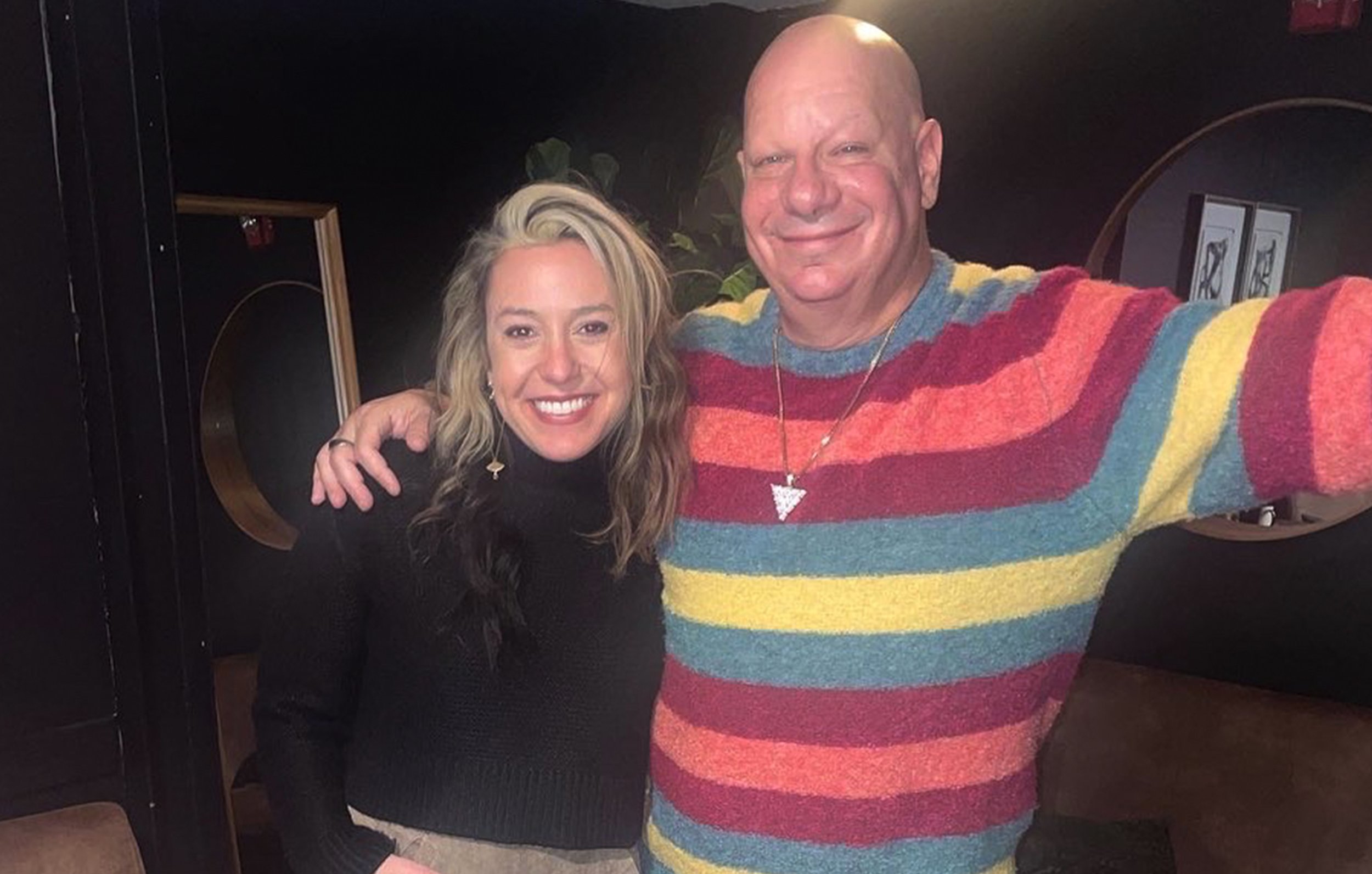 A man and woman smiling while taking a selfie together, with the man wearing a colorful striped sweater and the woman wearing a black top, in a dimly lit indoor setting.