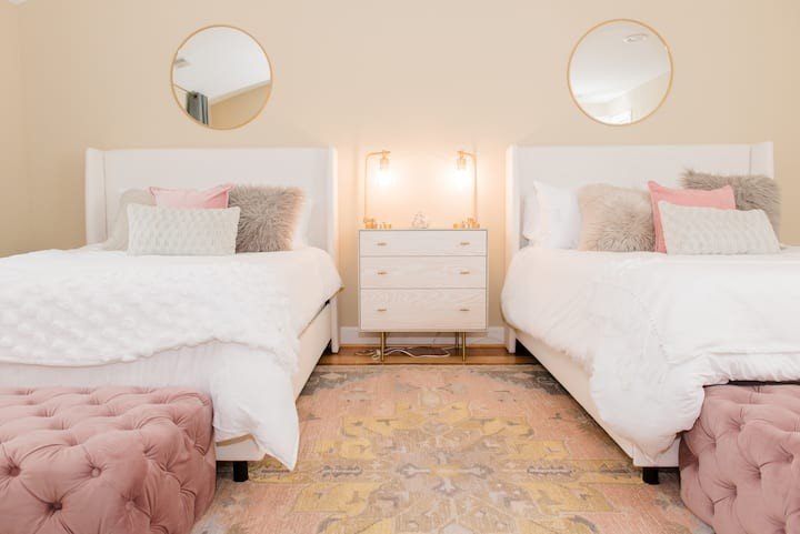 A bedroom with two white beds, each with pink and gray pillows, beige walls, a white dresser between the beds, and two round mirrors above the beds.