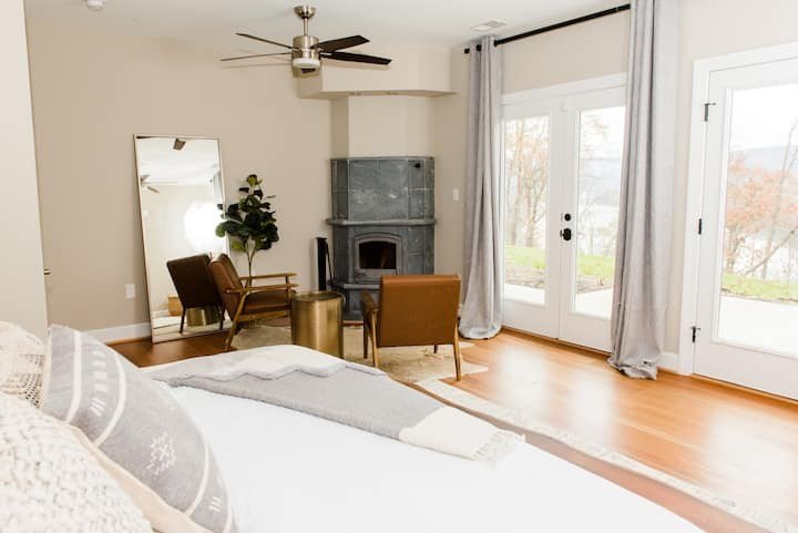 Cozy bedroom with bed, pillows, and blankets, a mirror, a potted plant, two chairs, a gold side table, a gray fireplace, and large glass doors with curtains leading outside.