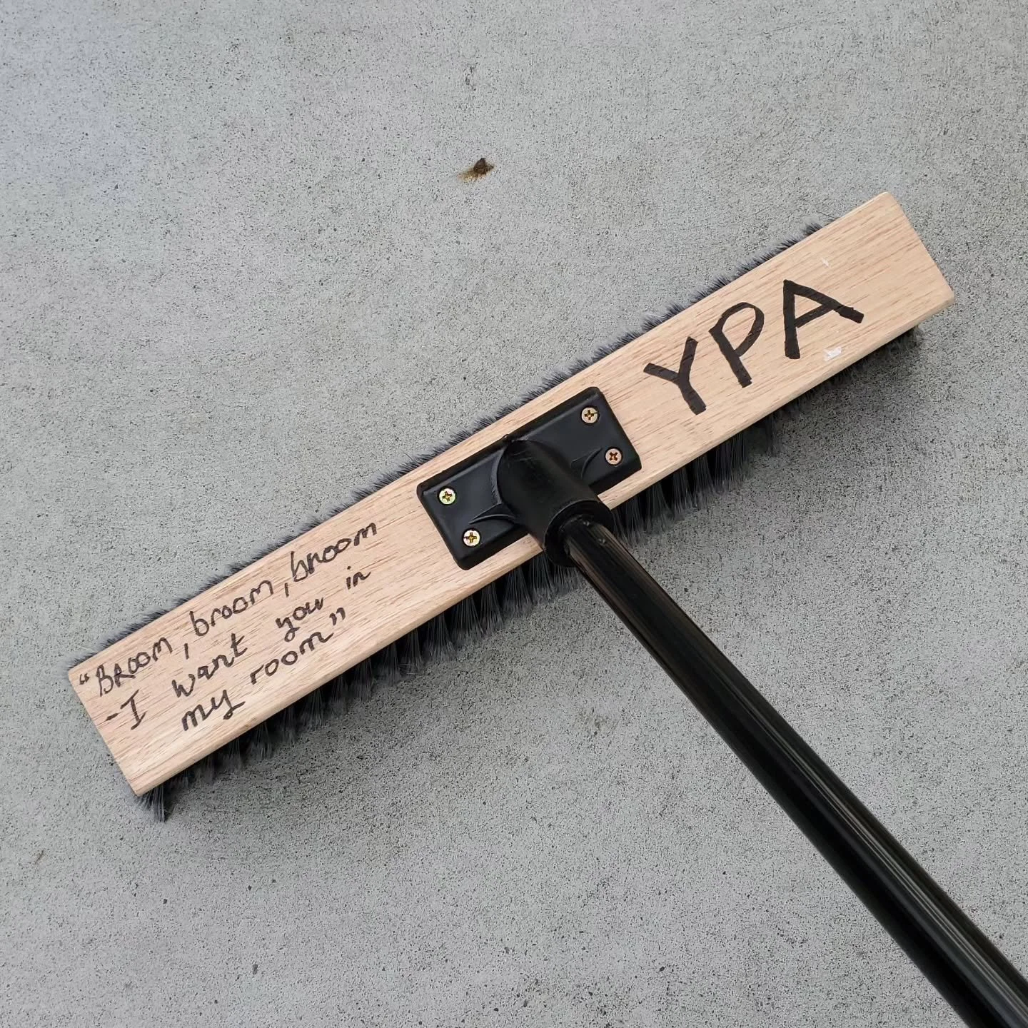 A long wooden brush with black bristles and a black handle. The word 'YPA' is written on the brush, and a quote is written on the handle that says, 'Broom, broom - I want you in my room'.