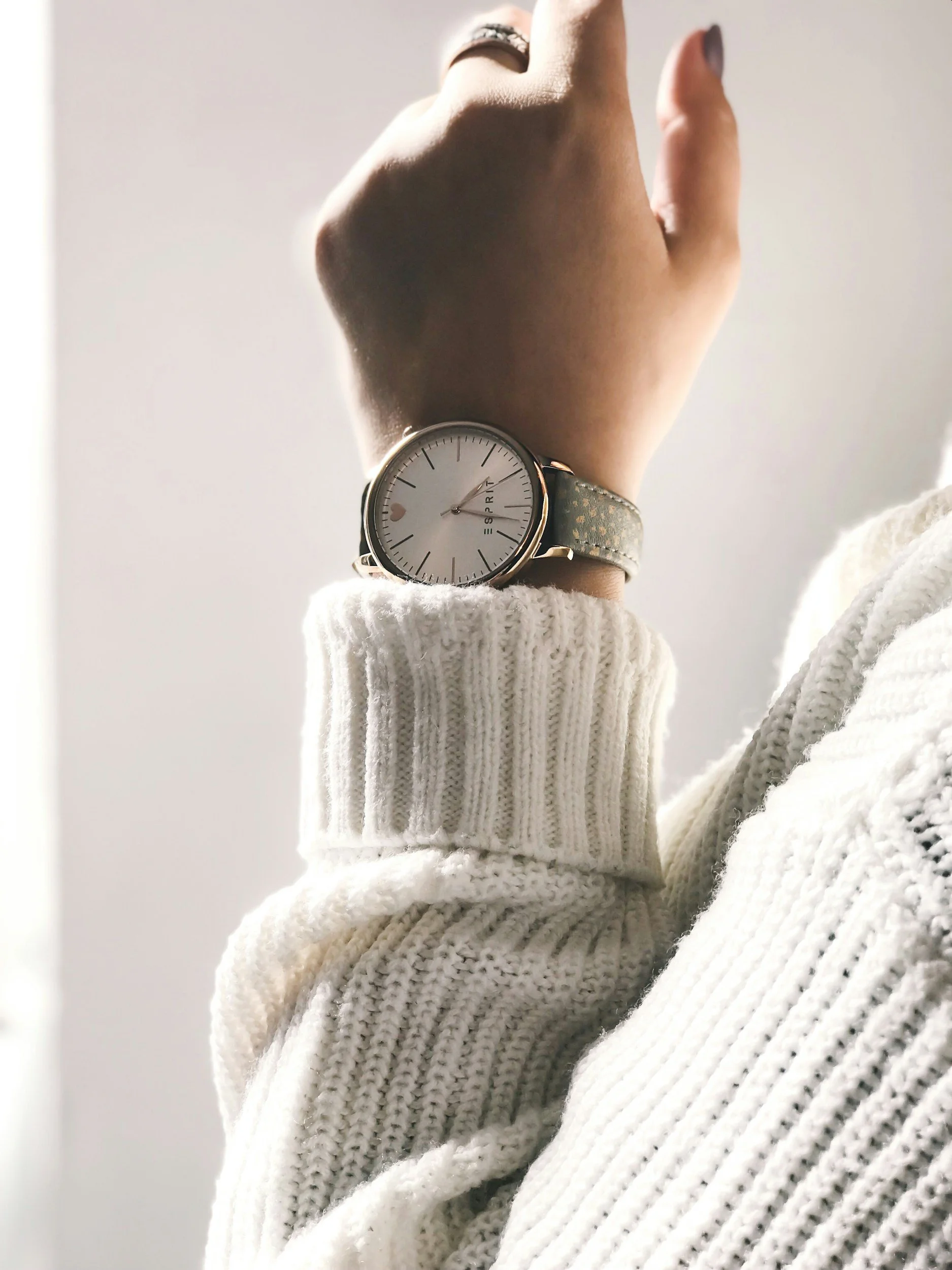 A person wearing a cream-colored knit sweater and a watch with a round face and a leather strap, showing a minimalist design with a white background and black markers, on their wrist.