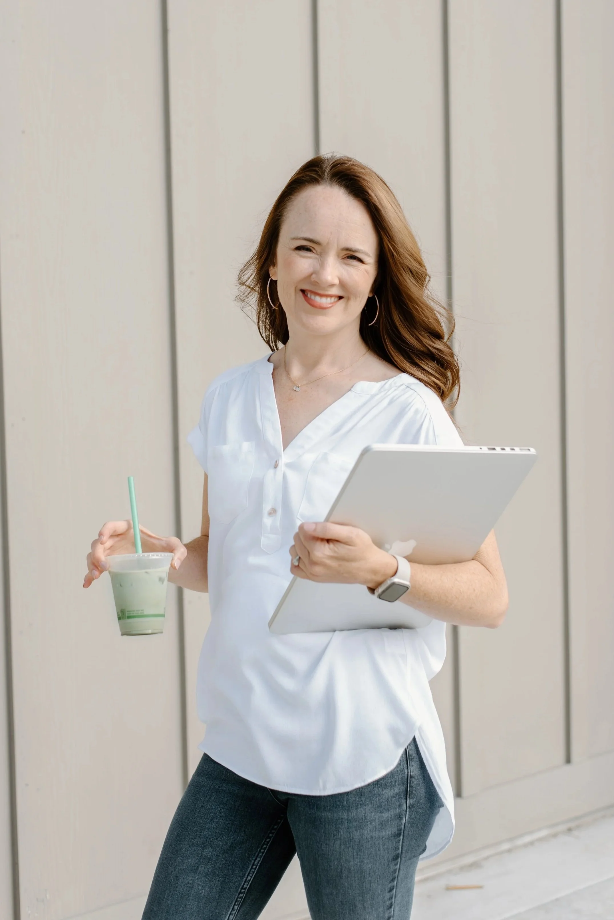 A woman with red hair smiling, holding a green smoothie and a silver laptop, standing outdoors against a beige wall.