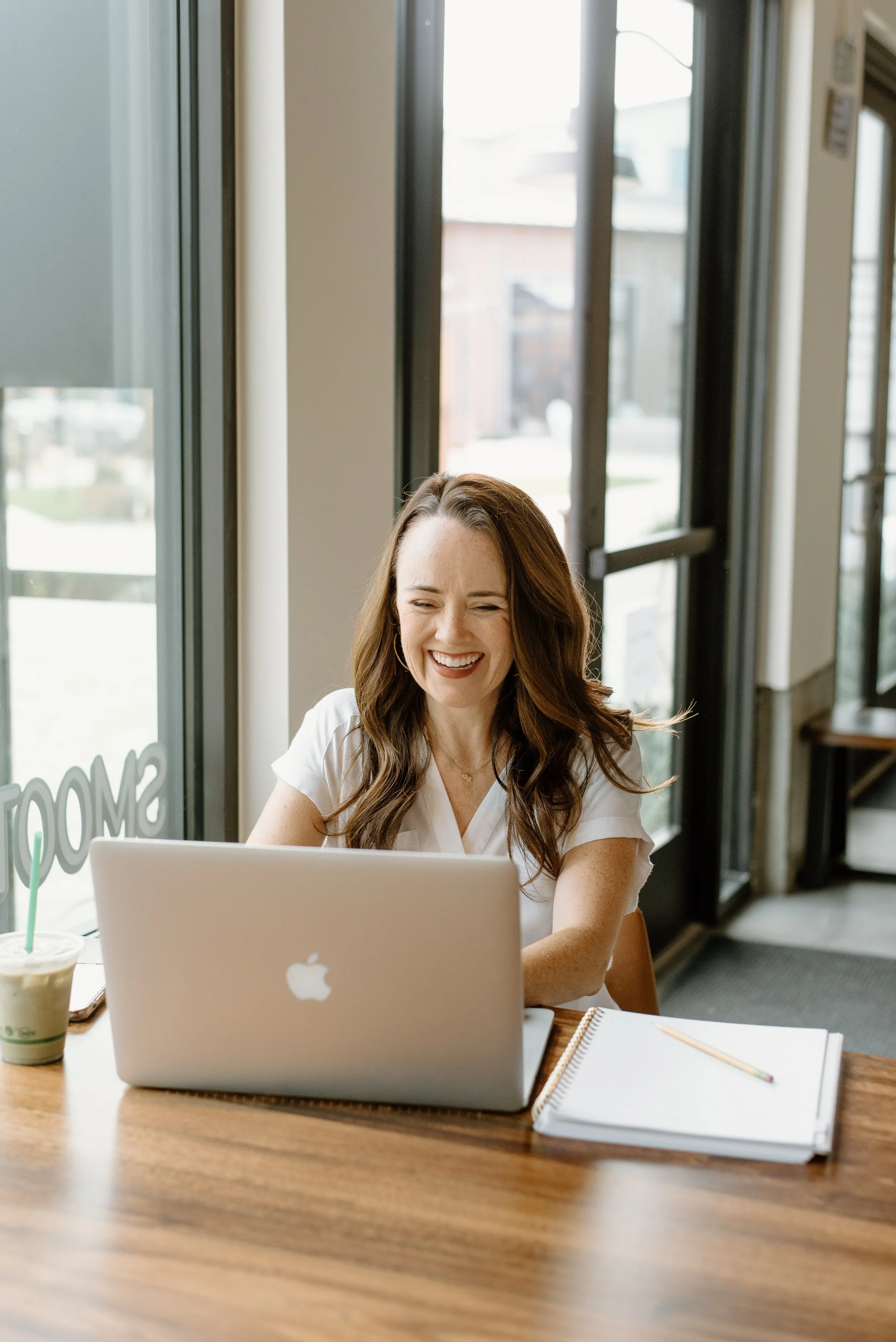 A woman with long brown hair, smiling and laughing while looking at her open MacBook laptop at a table in a cafe with large windows.