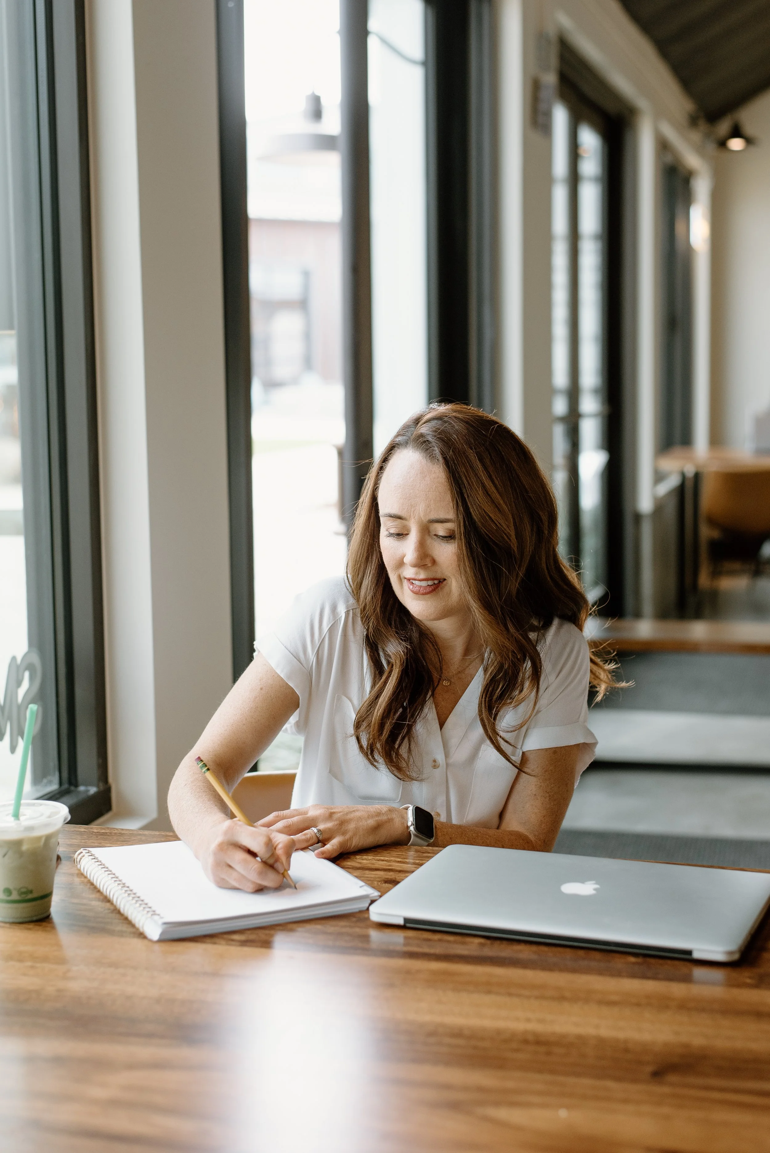 Woman with long brown hair sitting at a wooden table, writing in a notebook, with a MacBook and a green iced drink on the table in front of her, in a bright modern cafe or workspace with large windows.