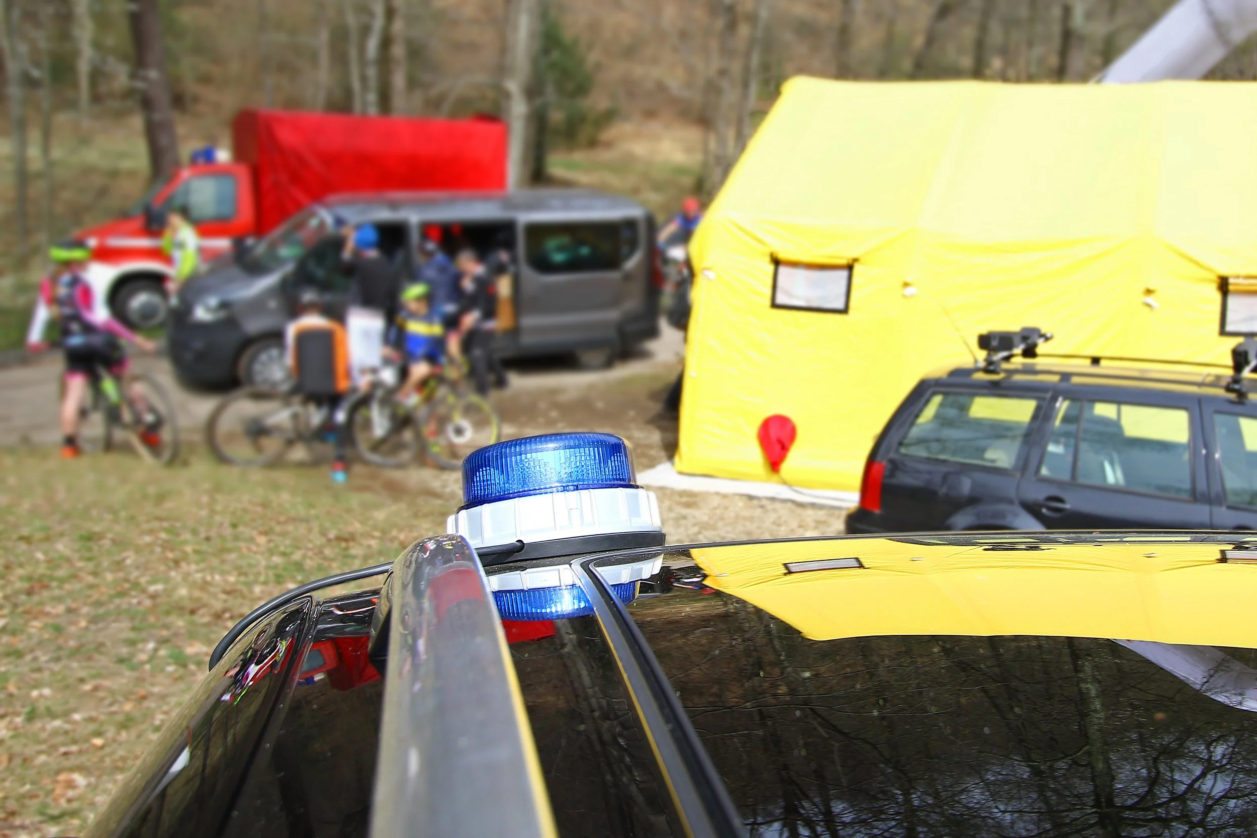 Emergency scene with police vehicle in foreground, blue light on roof, yellow tent, rescue workers, and cyclists in the background at an outdoor wooded area.