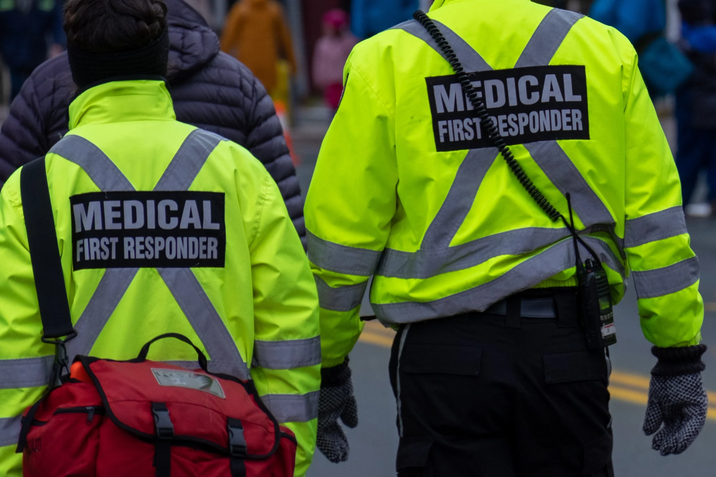 Two emergency medical first responders wearing bright yellow jackets with reflective stripes, walking in a crowd during what appears to be a public event.